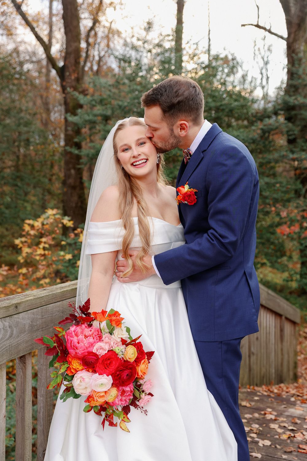 A joyful wedding moment captured on a wooden deck with autumn trees and a stunning pink and red bouquet.