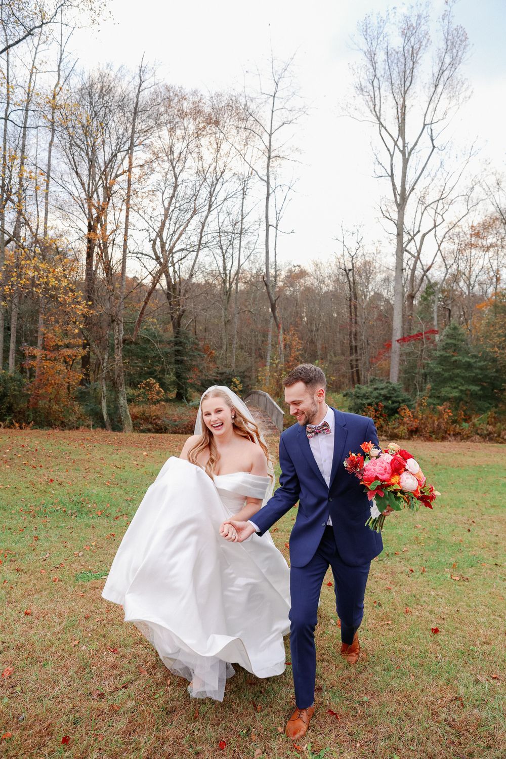 A couple in wedding attire playfully runs through an autumn field with fallen leaves and bare trees in the background.