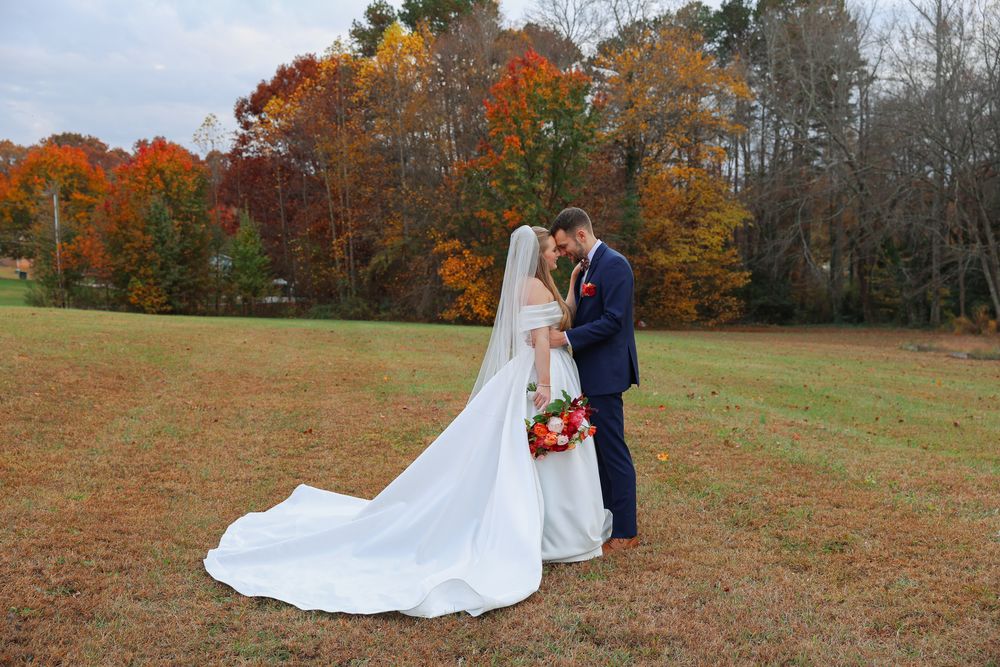 A couple in wedding attire share a romantic moment in a field surrounded by vibrant autumn foliage.