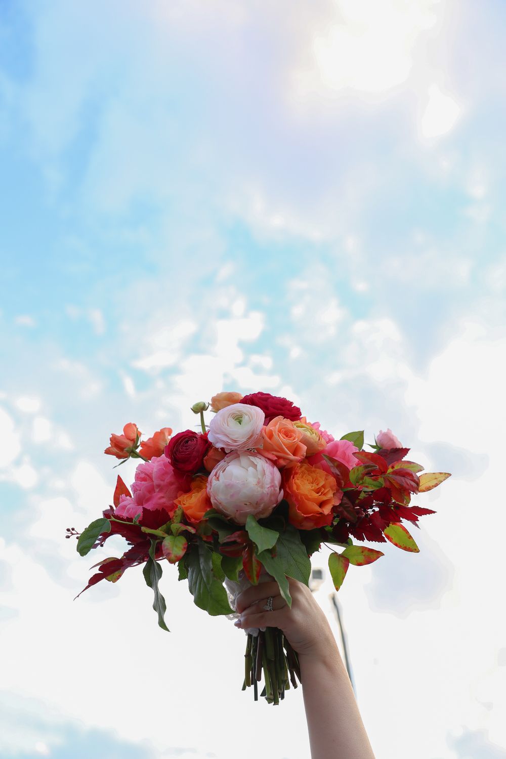 A colorful bridal bouquet with red, pink, and orange roses held up against a bright blue sky.
