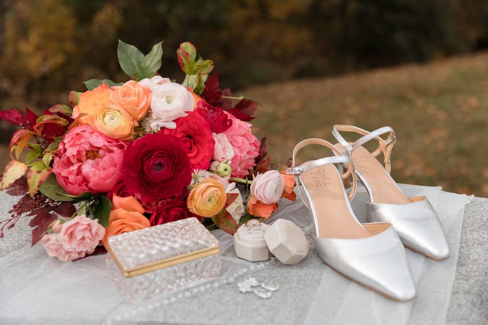 Wedding accessories including white shoes, pink and red floral bouquets, and delicate jewelry arranged on a rustic surface.