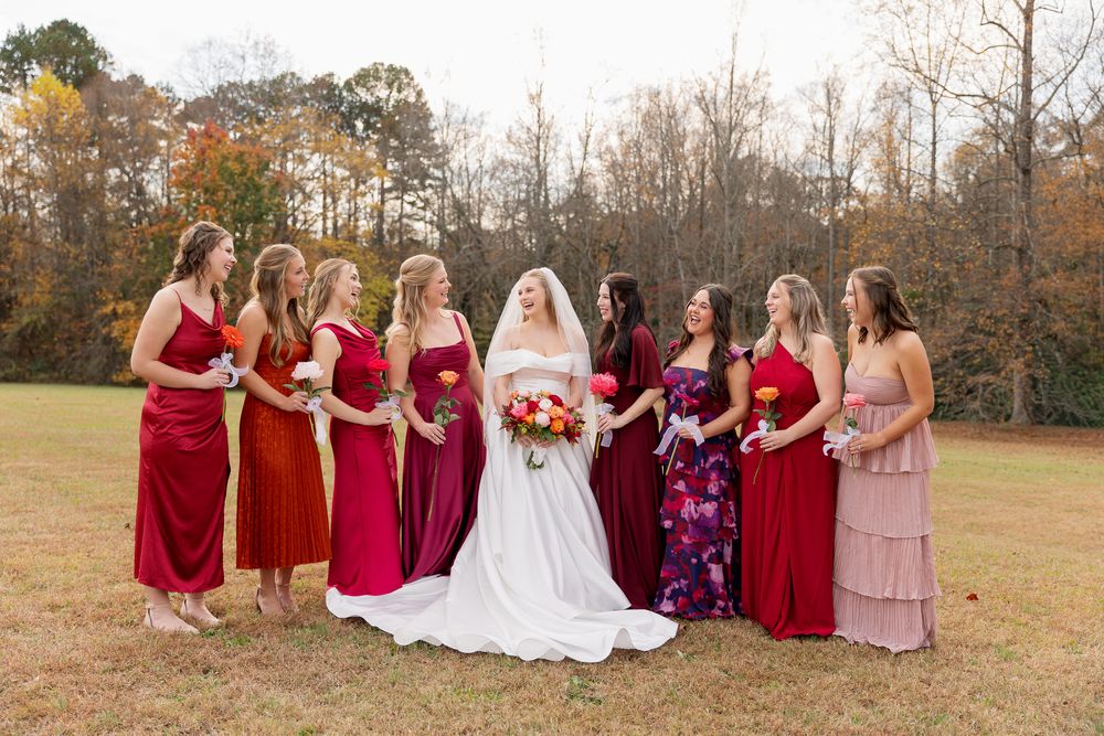 Bridesmaids in burgundy dresses sit together on a grassy field during an autumn wedding photoshoot.