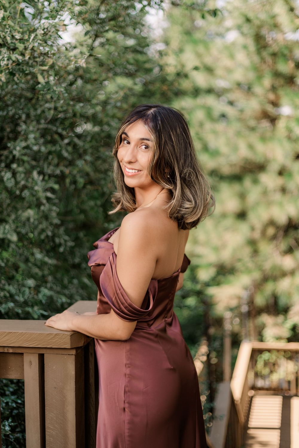 Woman in a burgundy dress poses on a wooden deck surrounded by lush greenery.