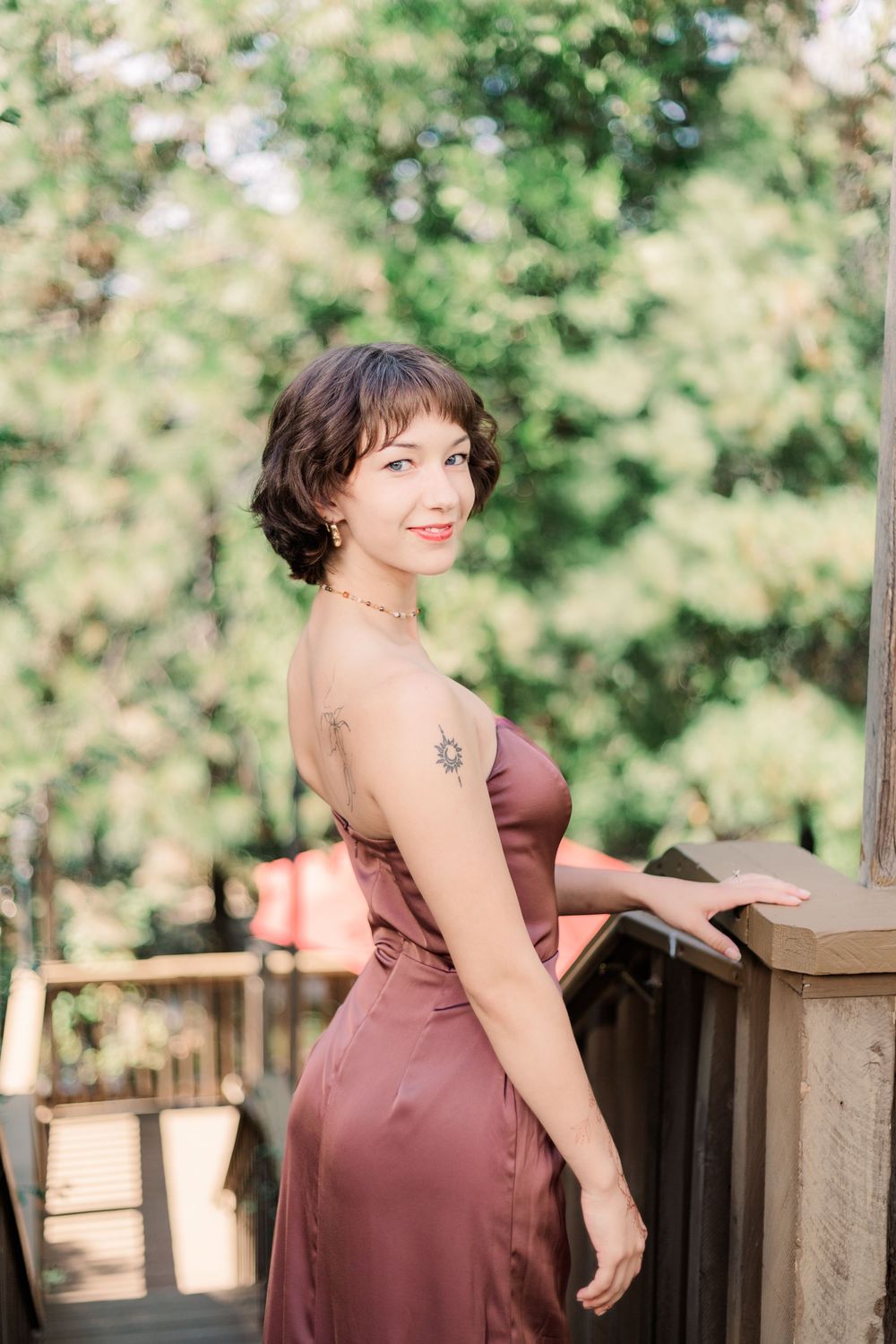 Woman in mauve satin dress poses on wooden deck railing with green foliage in background.