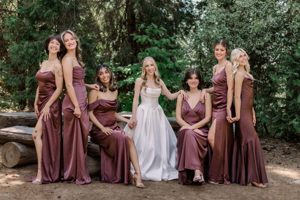 Wedding party wearing brown dresses poses together outdoors in a garden setting with a person in white.