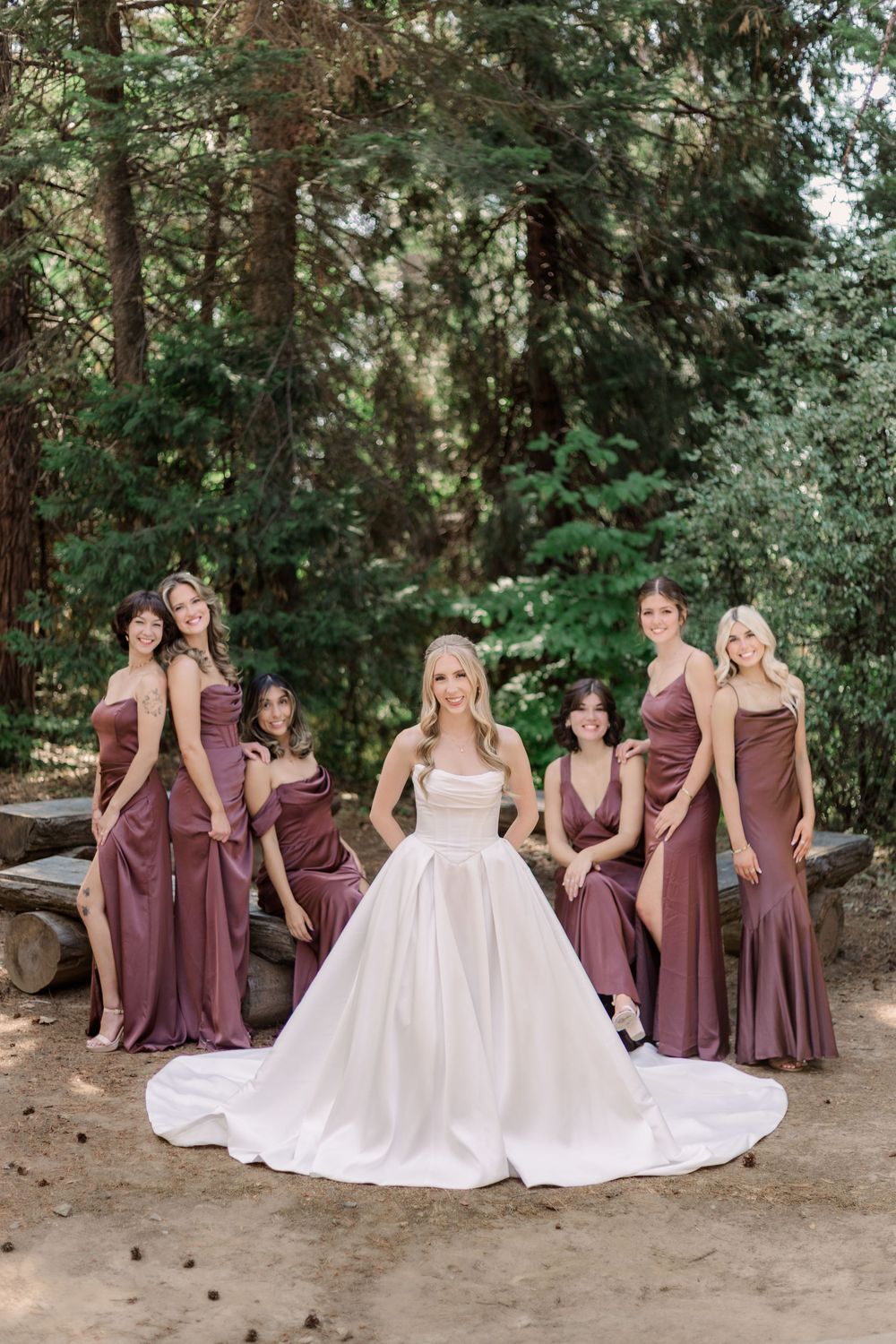 Wedding party poses in a forest setting with bridesmaids in burgundy dresses surrounding bride in white gown.