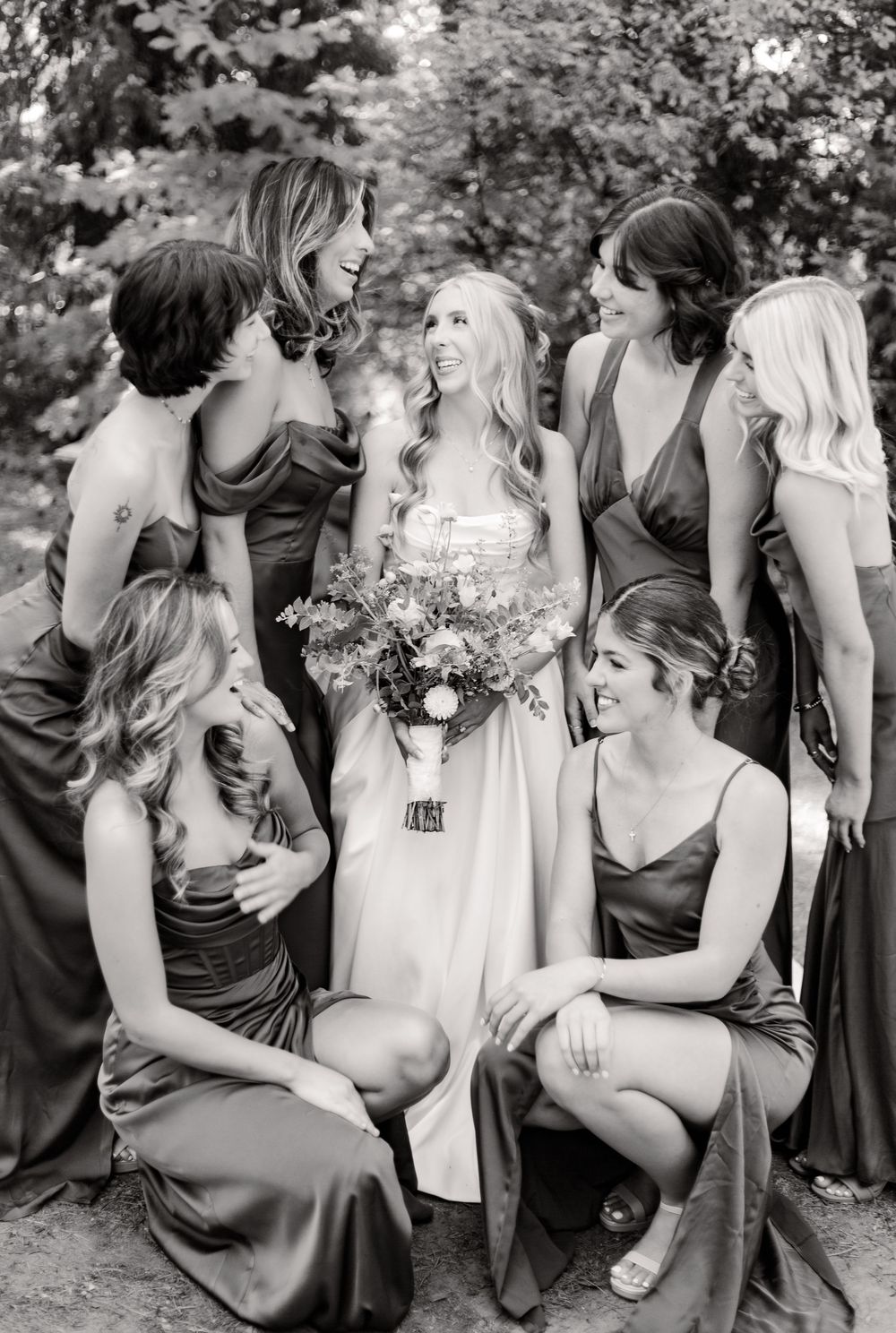 A group of bridesmaids in brown dresses sit together outdoors for candid black and white wedding photos.