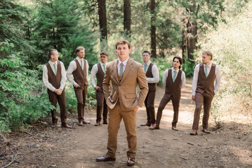 Wedding party members in brown suits gather outdoors in a wooded setting for candid photos.