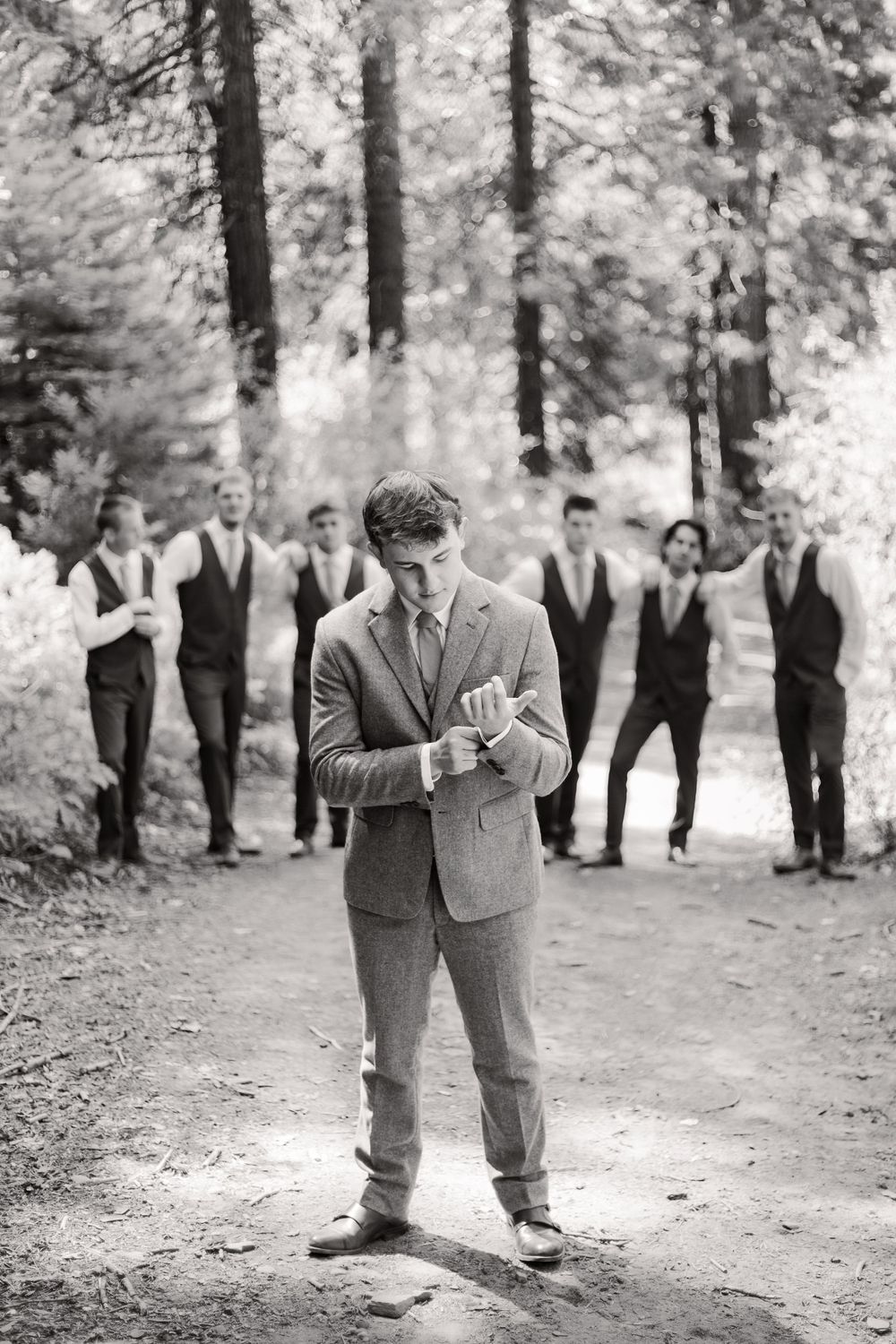 Black and white photos show groomsmen in suits sharing playful moments in a wooded outdoor setting.