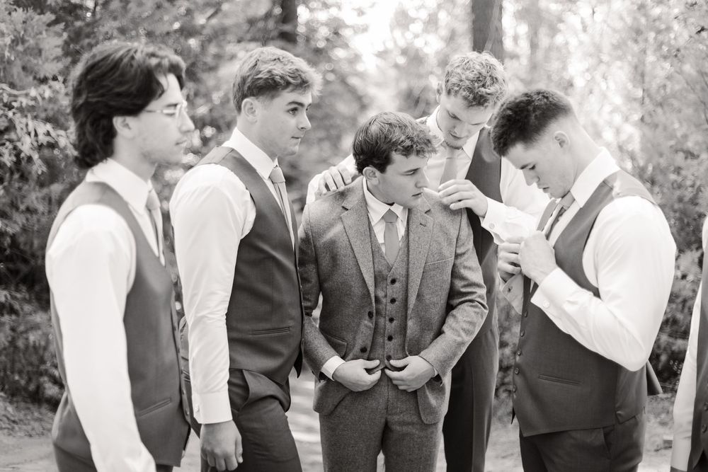 Groomsmen in formal attire interact during an outdoor wedding celebration in a forest setting.