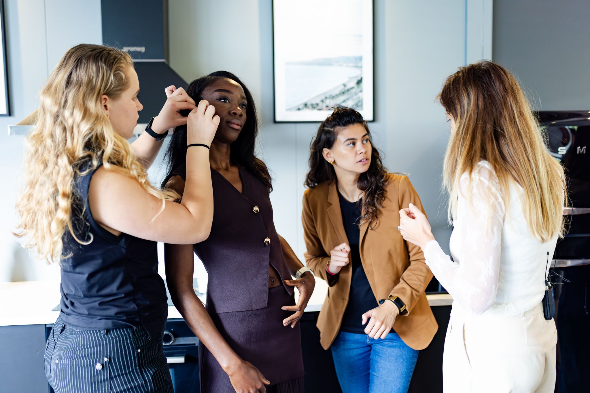 A group of women laugh together during a makeup session in a bright modern office space.