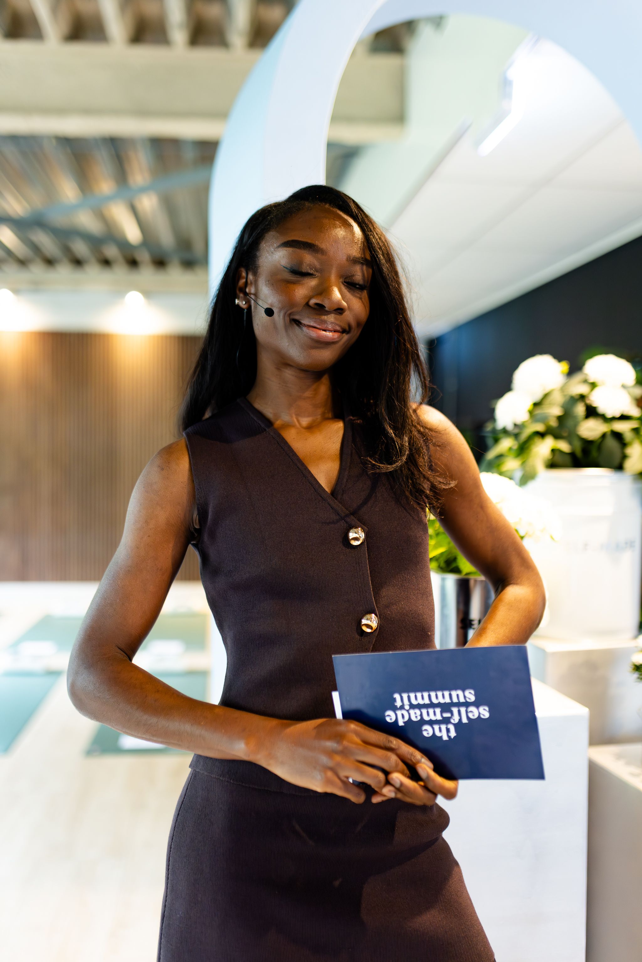 A woman in a brown dress poses with a small blue booklet in a modern office setting.