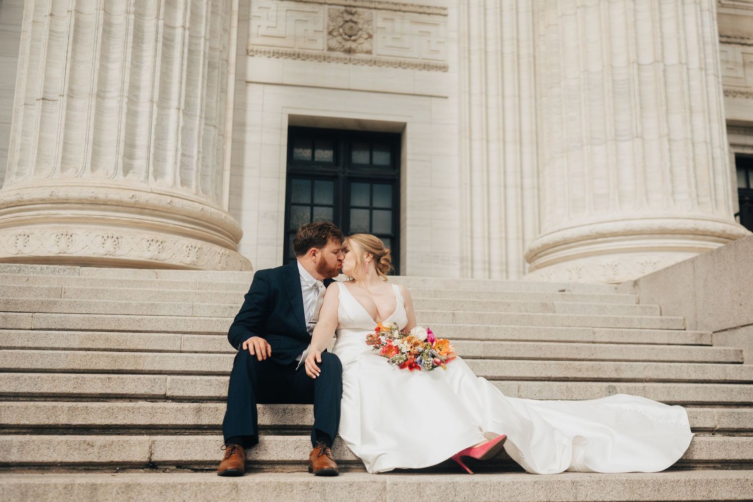 Bride and groom sharing a romantic kiss on stone steps of a grand columned building on wedding day.