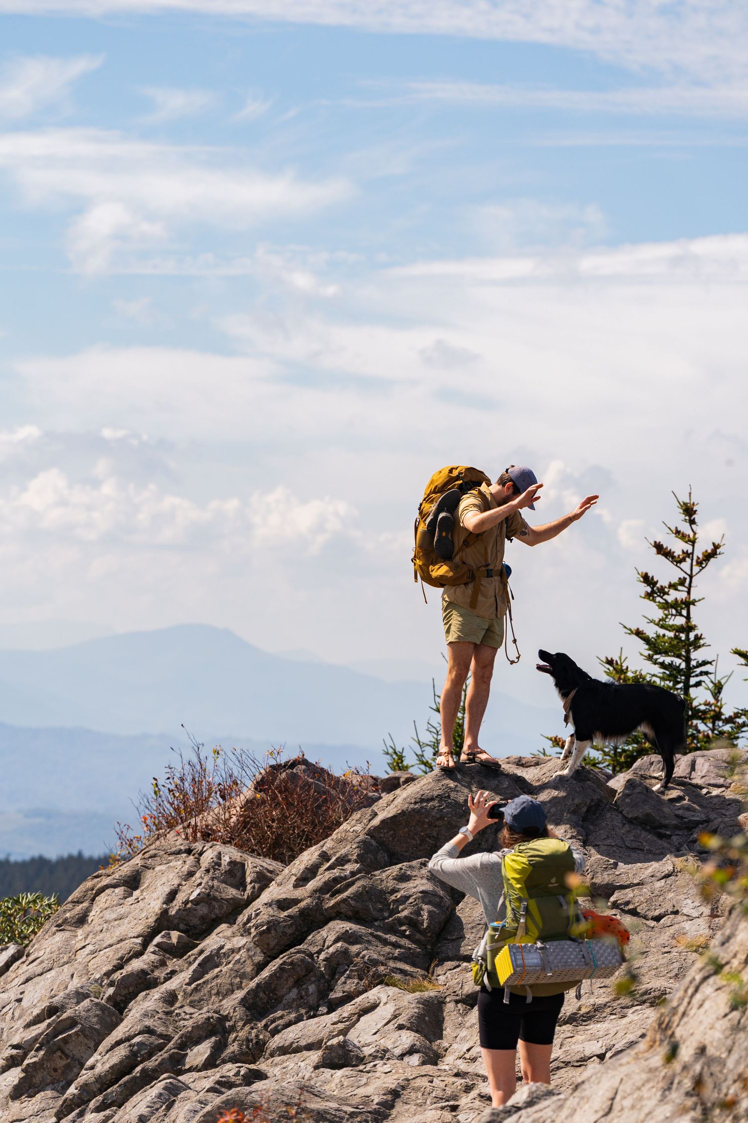 Hikers with backpacks traverse rocky mountain terrain under blue skies with distant peaks in view.