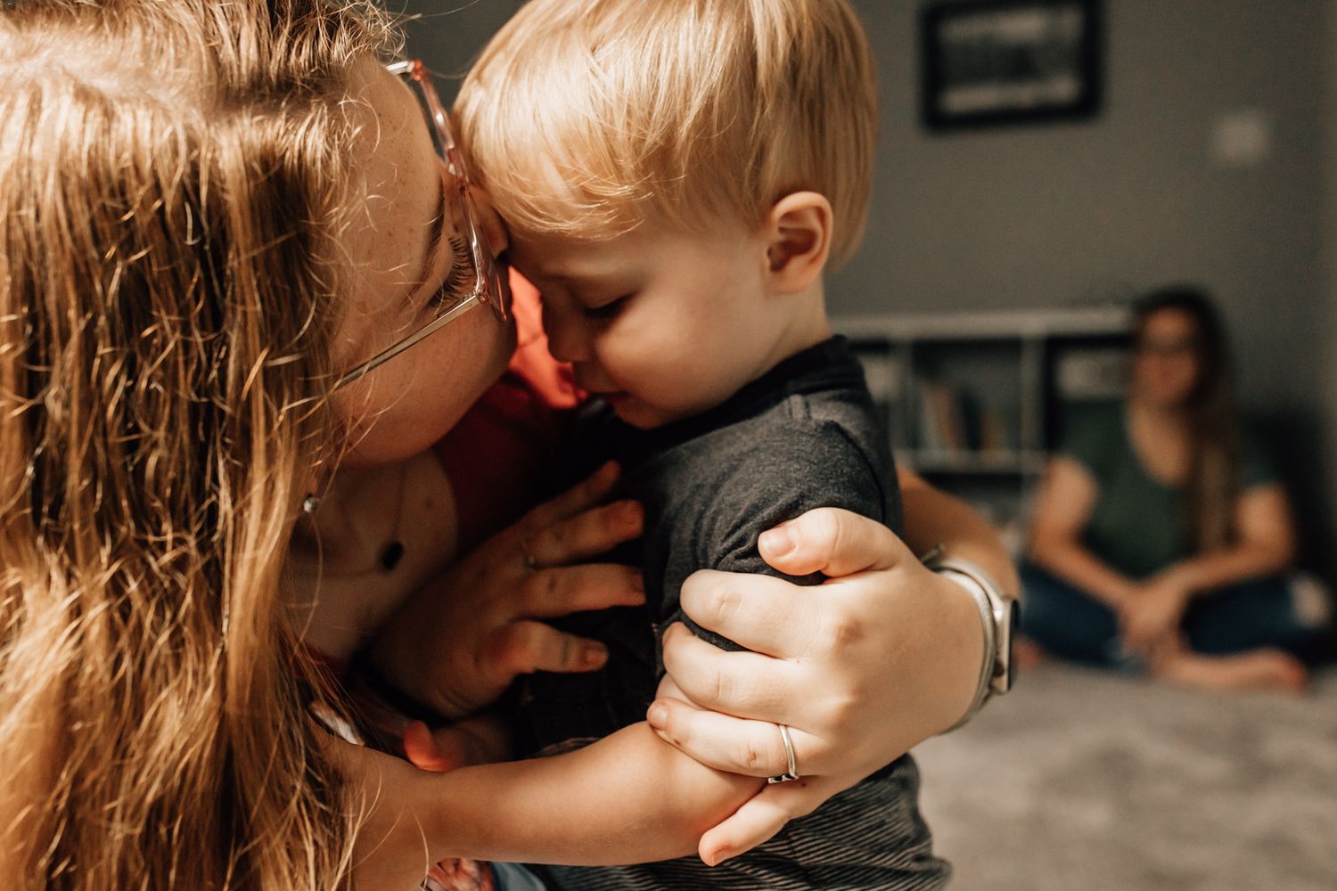 A tender moment between half sister and brother sharing a sweet embrace in a dimly lit room with mom seated in background. Whitney Popp