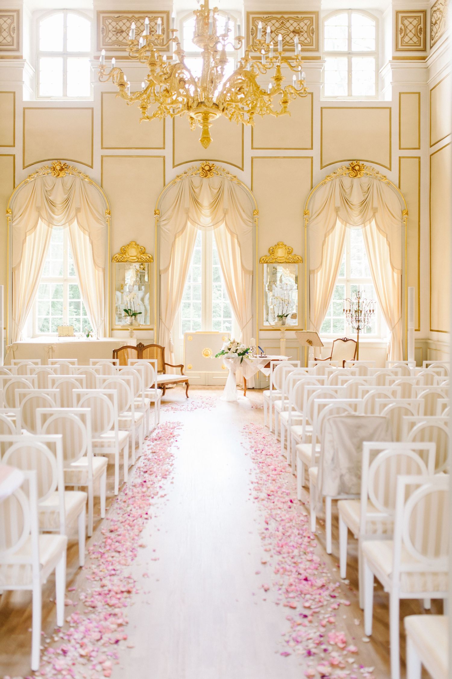 Elegant wedding ceremony setup with white chairs, pink rose petals along the aisle, and ornate gold chandelier in a luxurious ballroom.