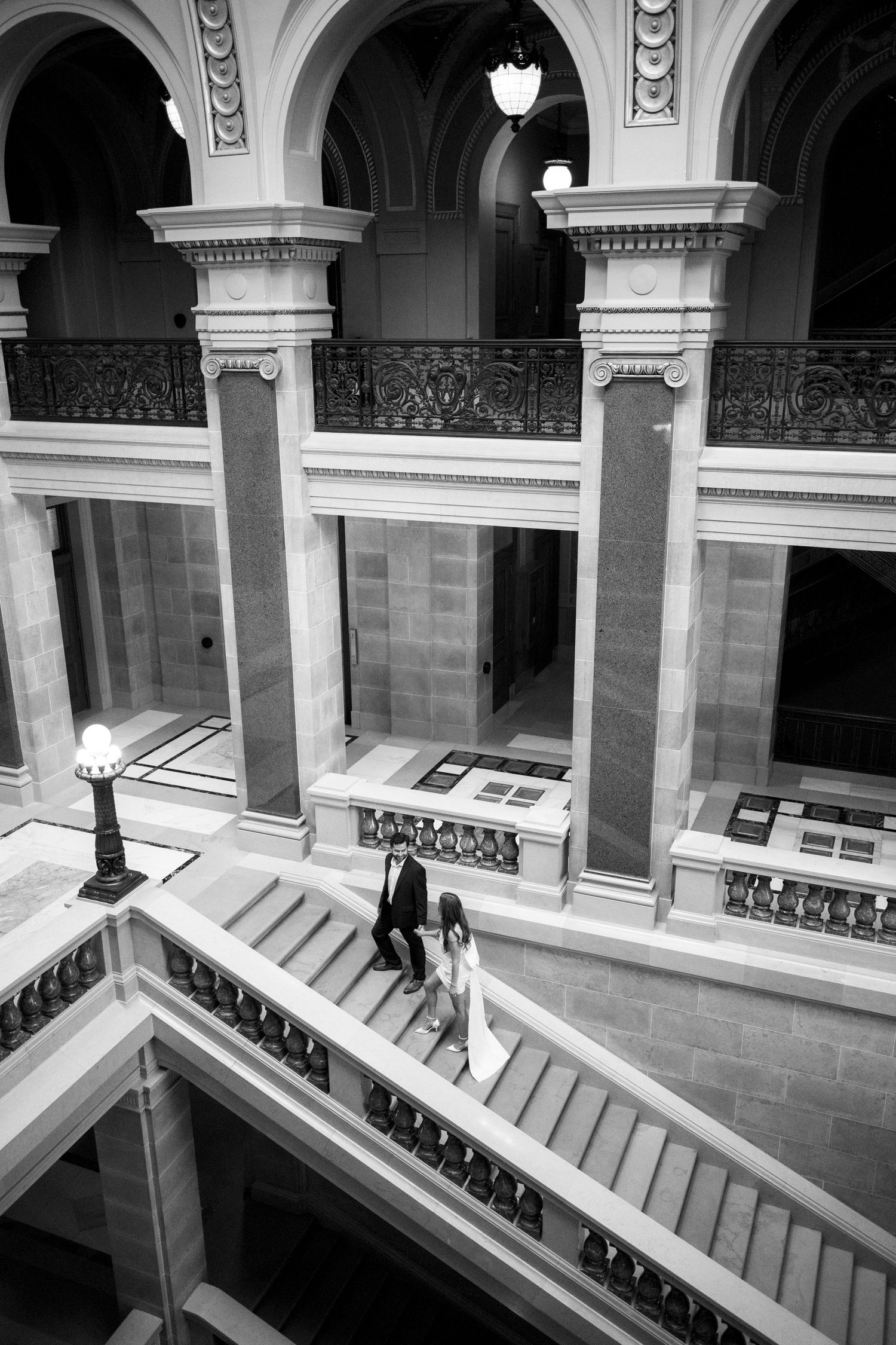 A couple descends an ornate marble staircase in a historic building with classical architecture.