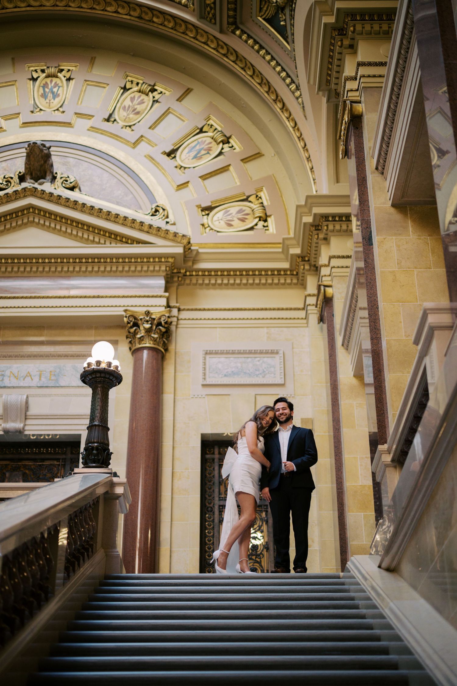 A couple poses on grand marble stairs inside a historic building with ornate architectural details.