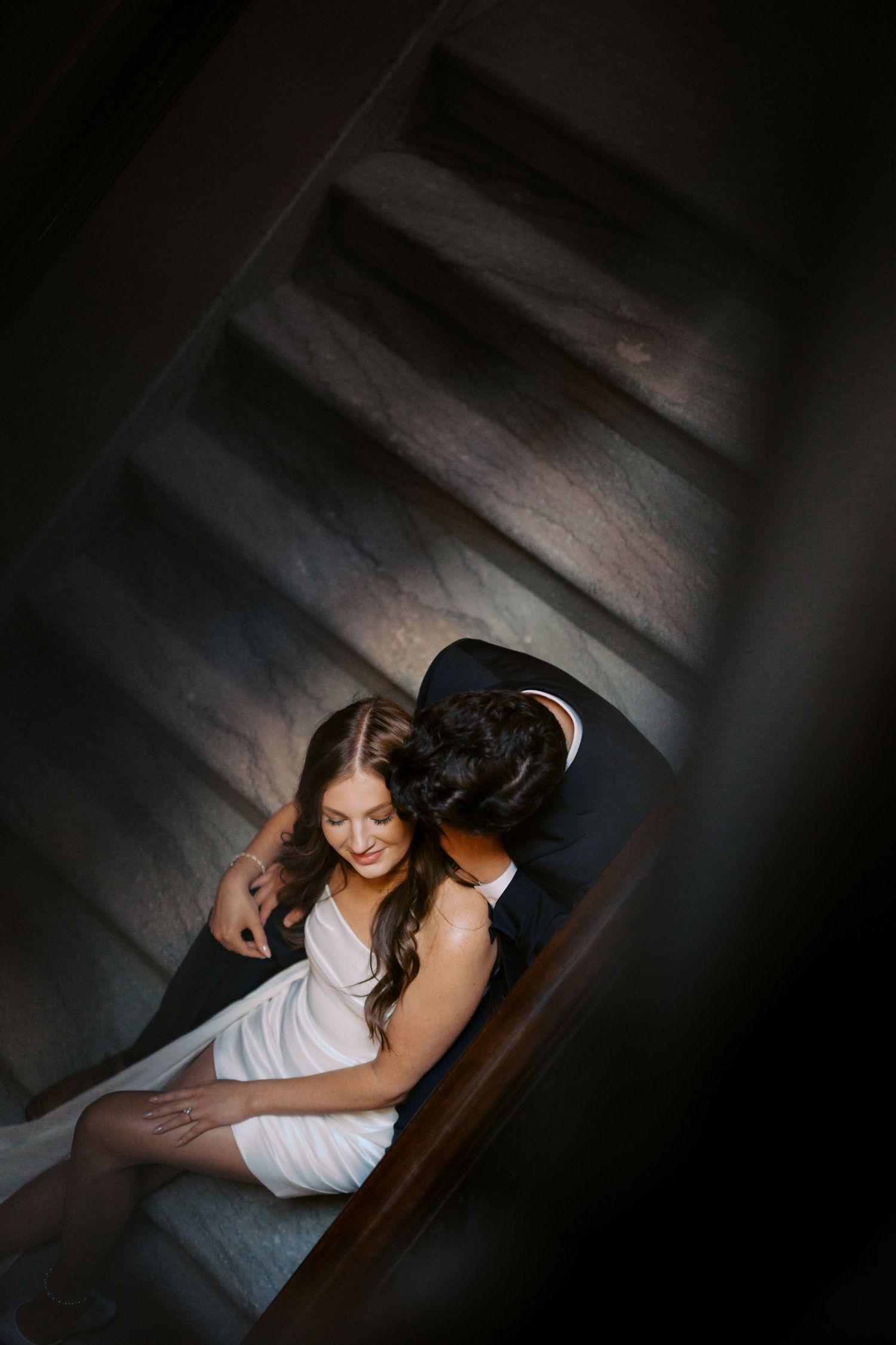 An overhead view of a couple sitting close together on dark wooden stairs.