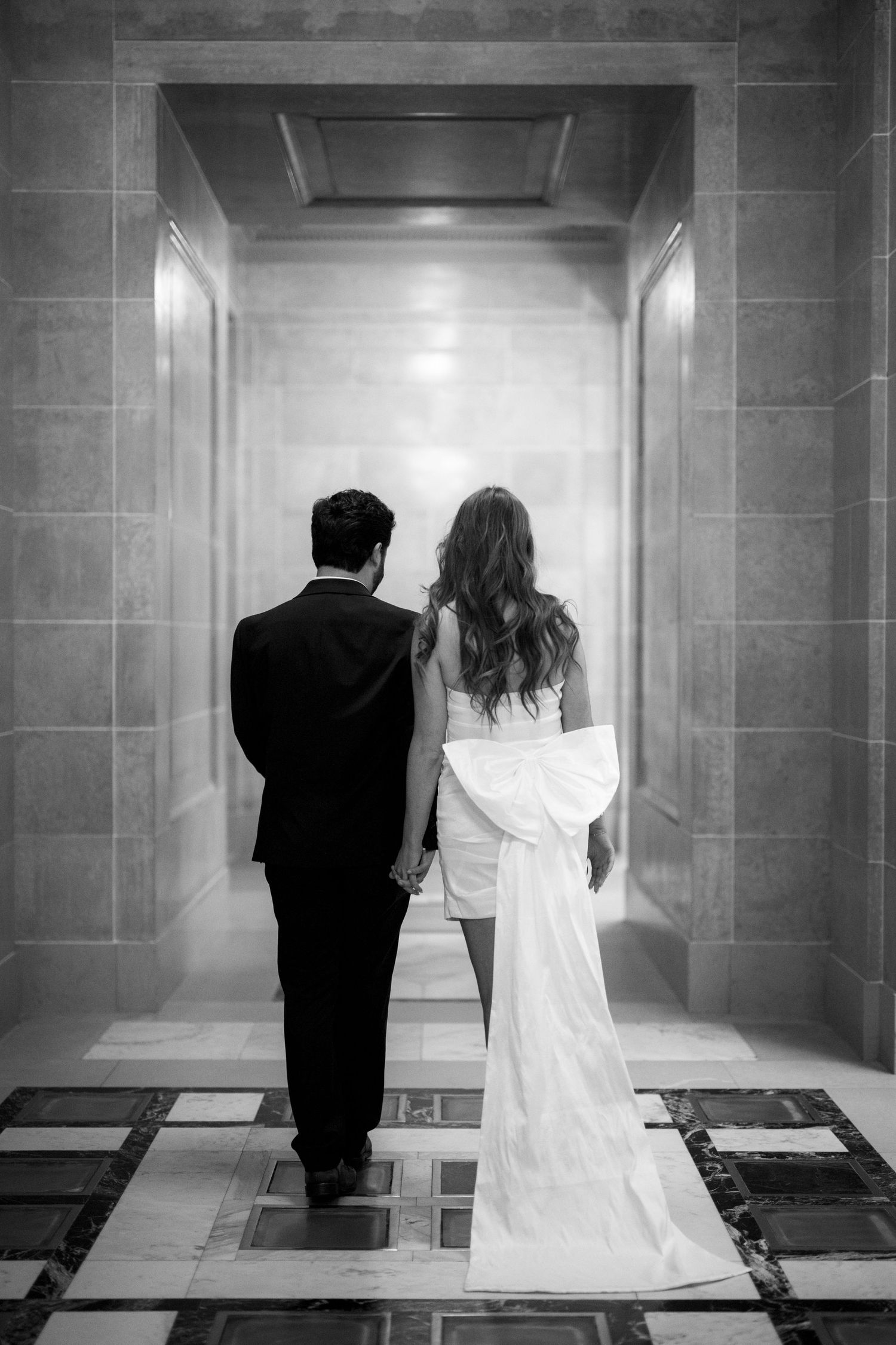 A couple in formal attire stands in an elegant marble hallway viewed from behind.