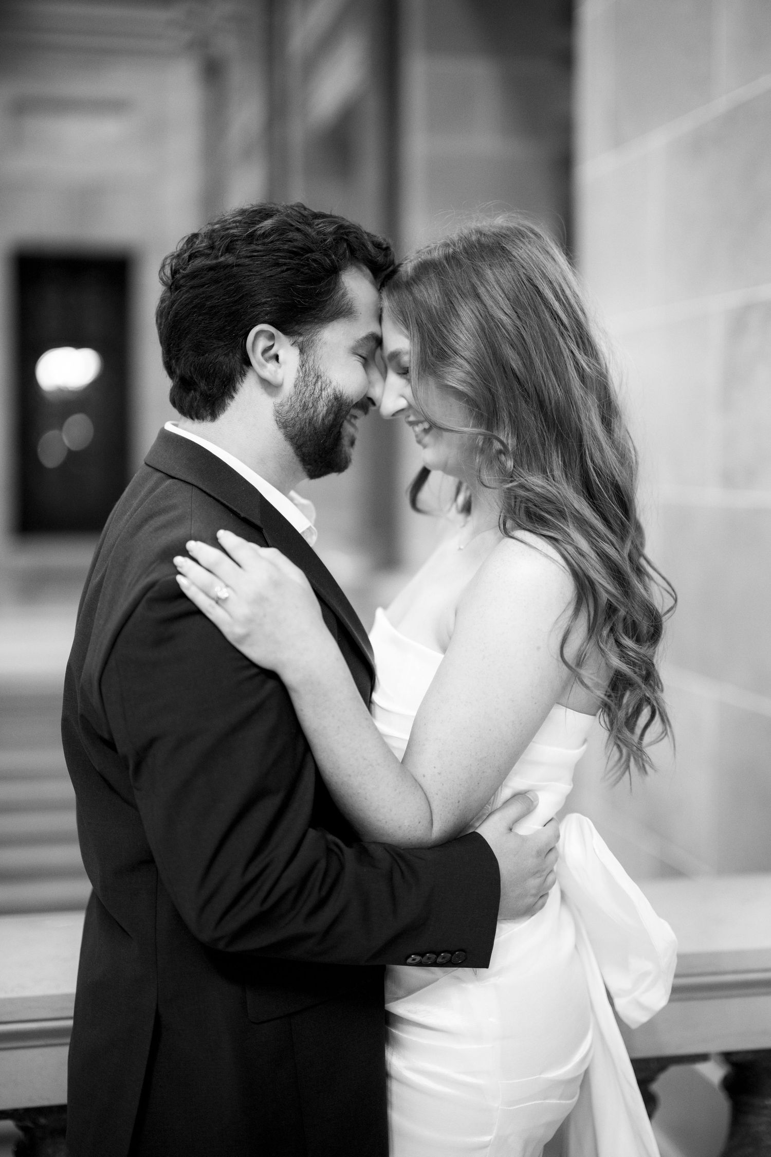 A romantic black and white portrait of a couple embracing in formal attire against a classic architectural backdrop.