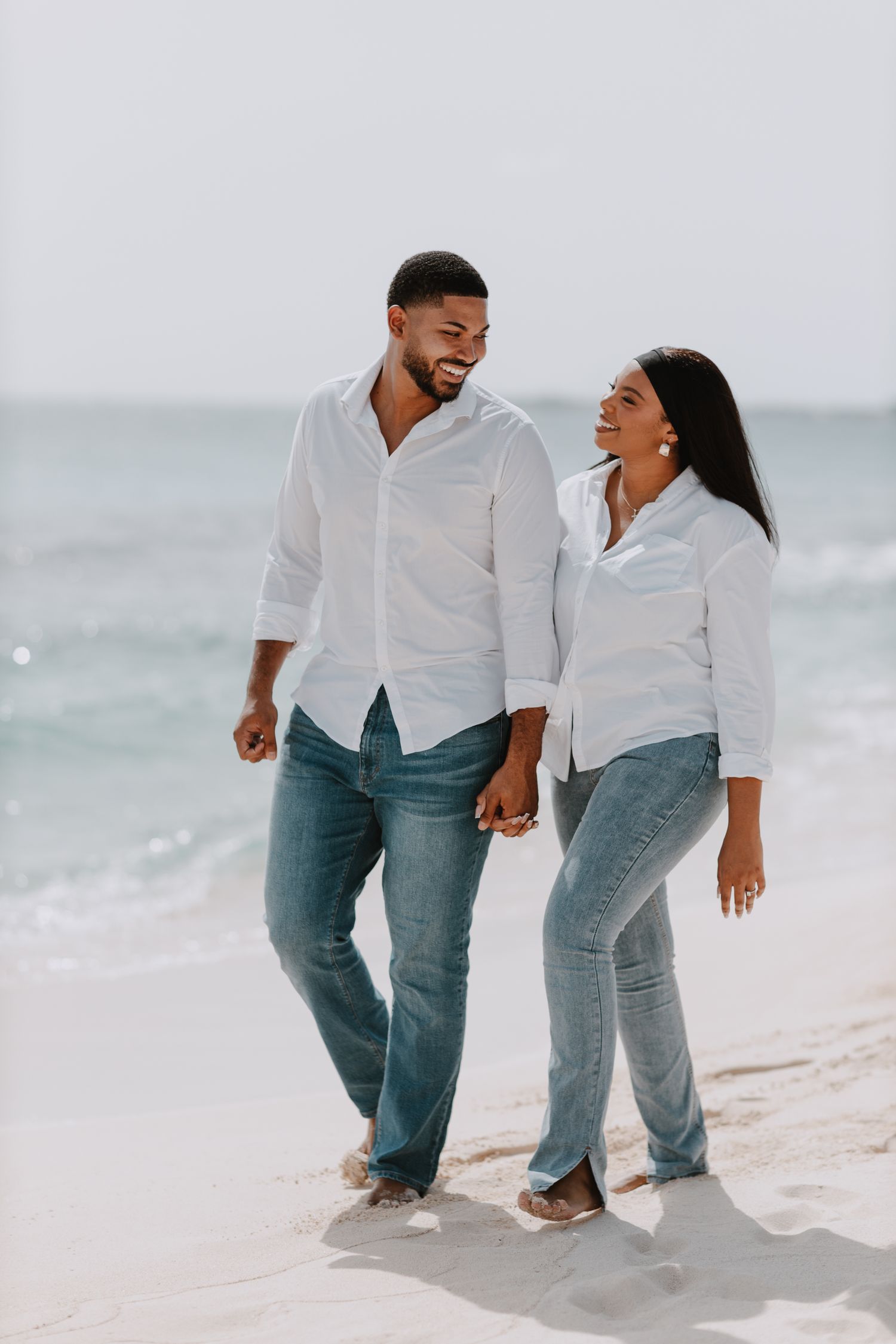 Couple wearing matching white shirts and jeans walking together on a sunny beach.