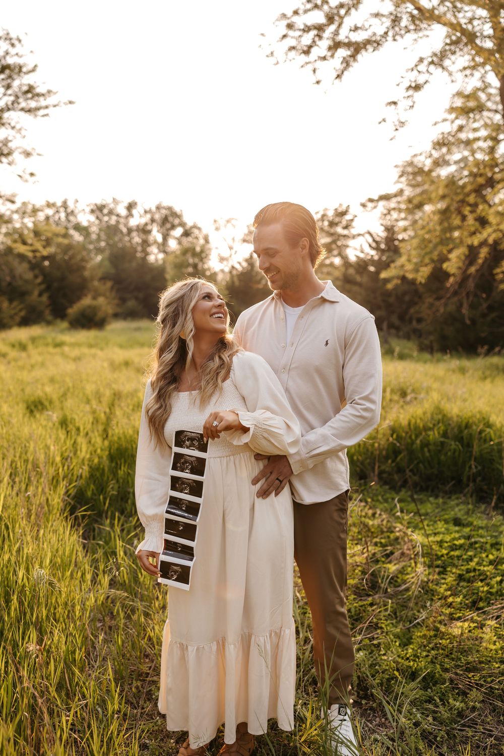 Joyful couple holding ultrasound photos while embracing in sunlit meadow.
