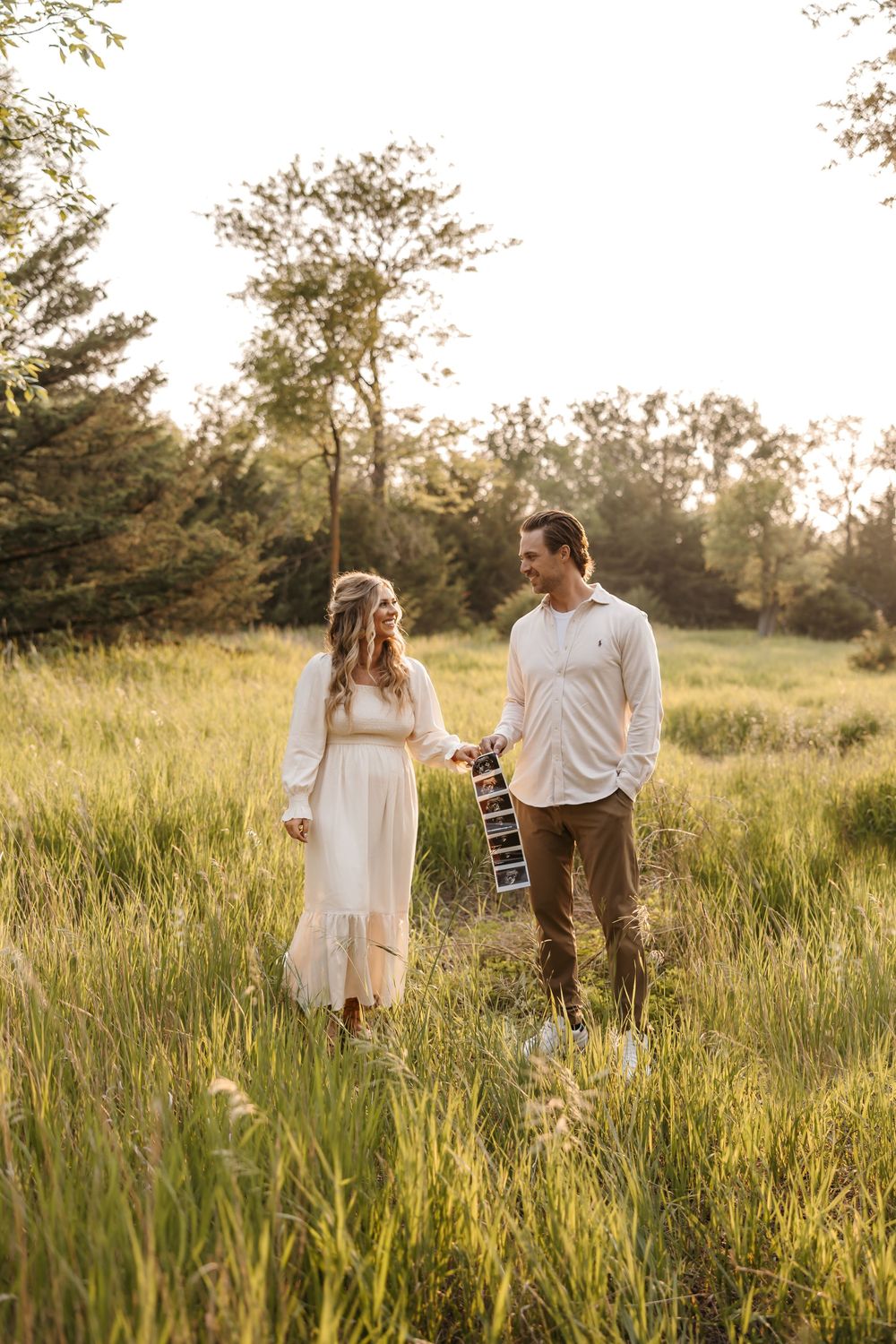 Couple walking through tall grass field at sunset holding ultrasound photos.