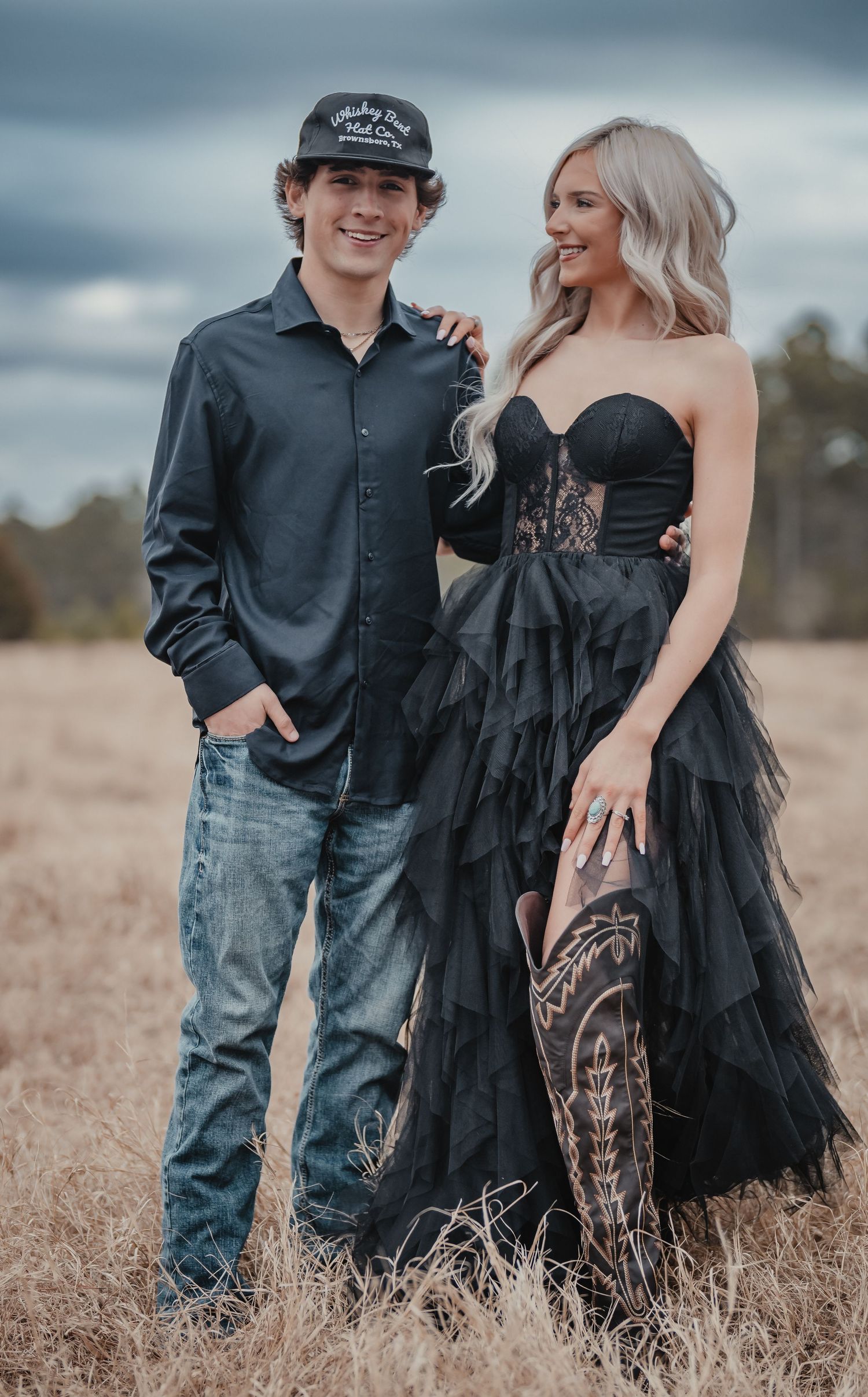 Couple posing together in a field with the woman wearing an elegant black strapless gown with layered tulle details.