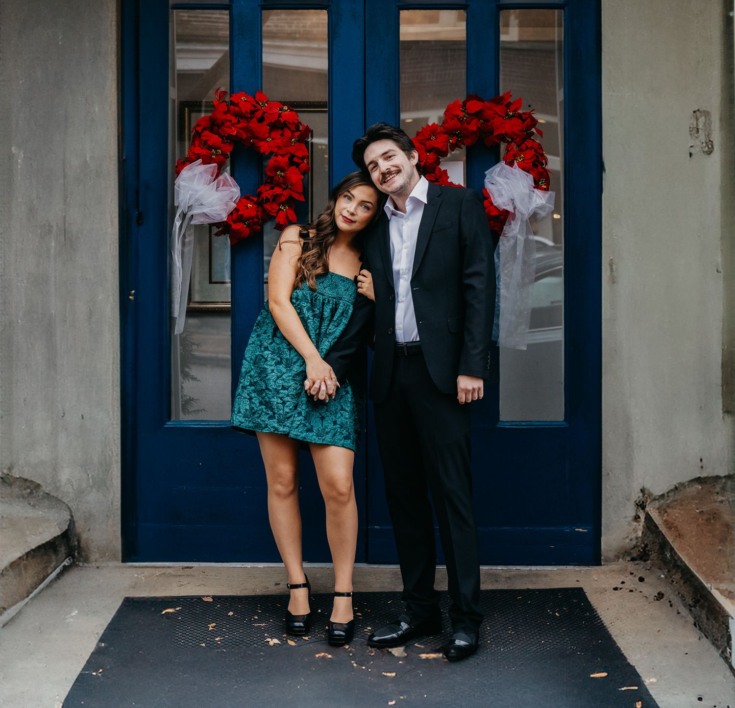 Couple posing in formal attire in front of a blue door decorated with red holiday wreaths.