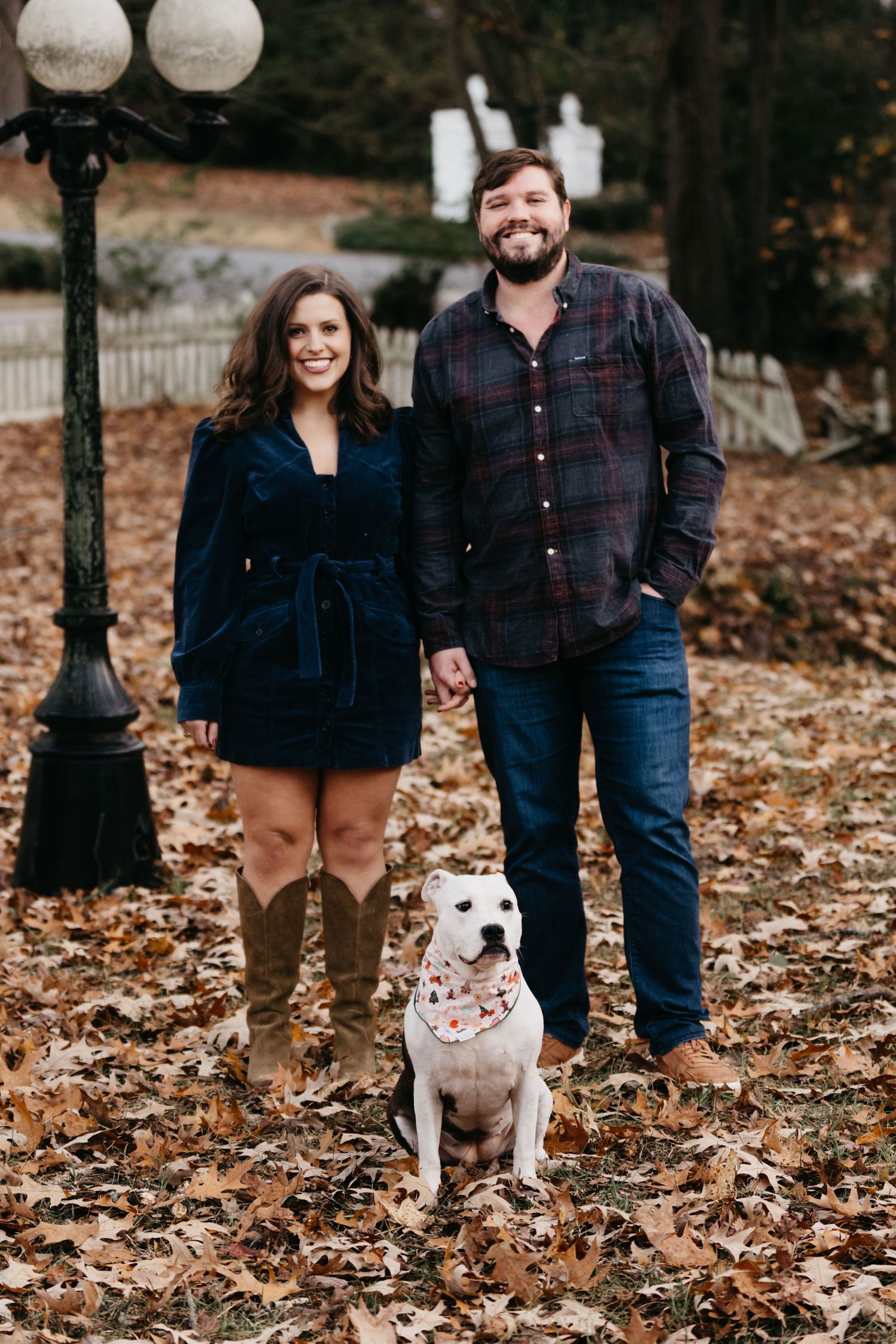 Couple and white dog pose together by vintage lamp post on autumn path.