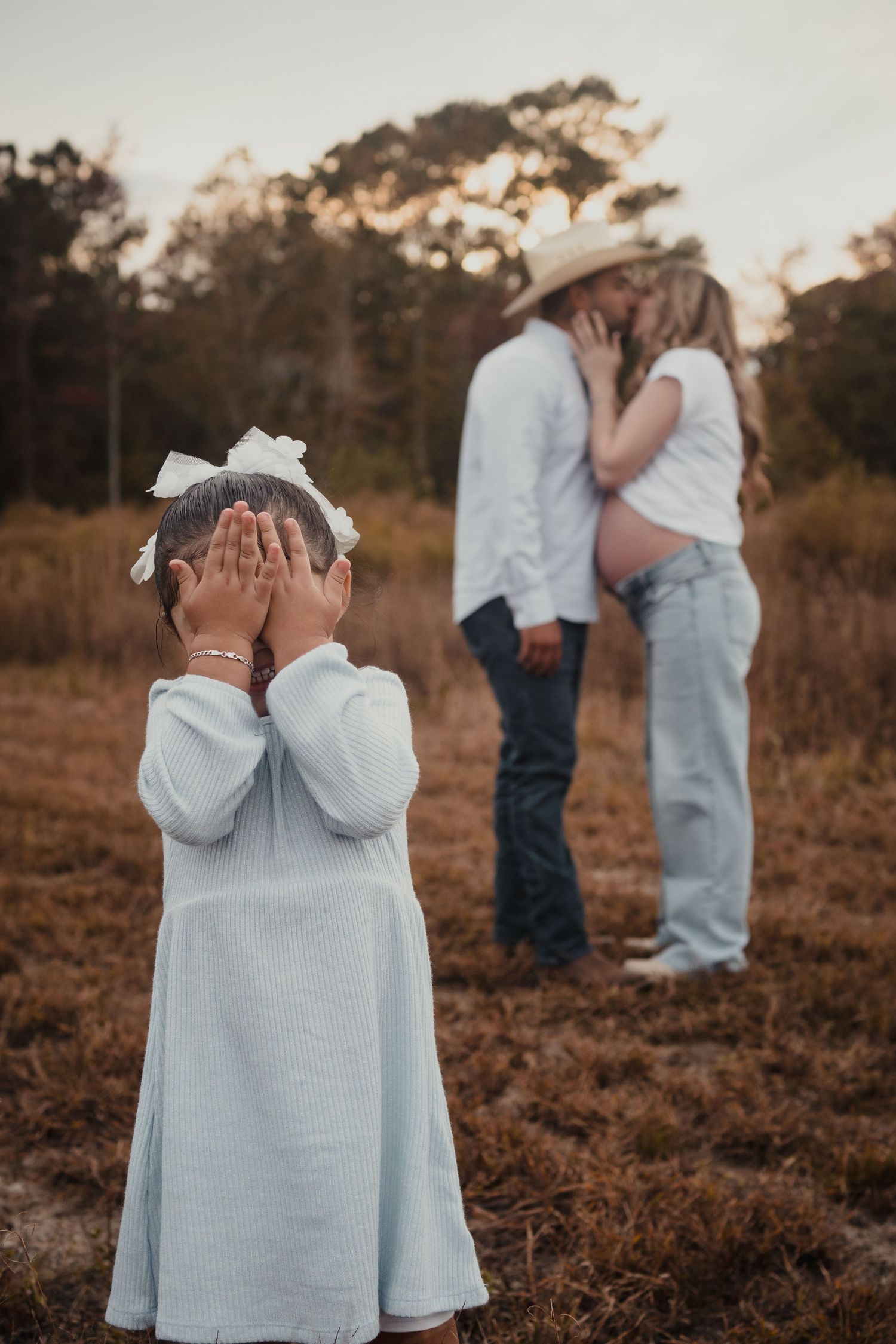 A child in a light blue sweater covers their face with their hands while playing peek-a-boo outdoors at sunset.