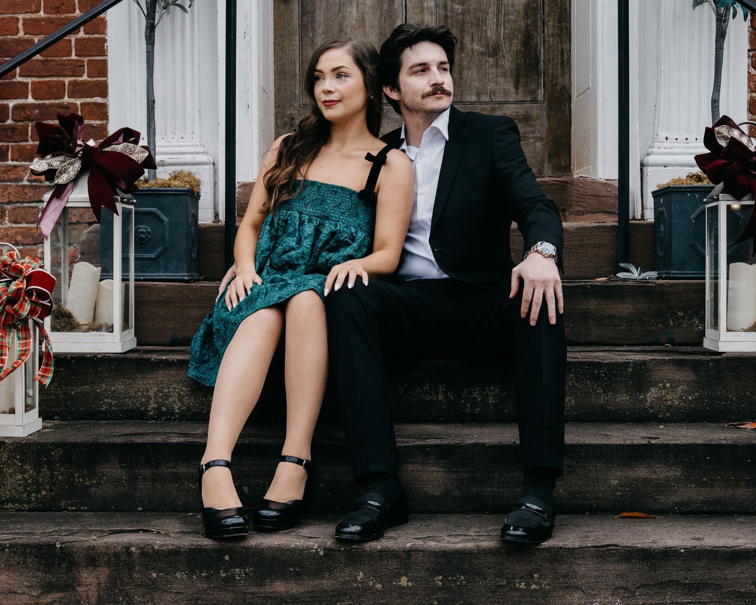 Couple in formal attire sitting on stone steps with holiday decorations.