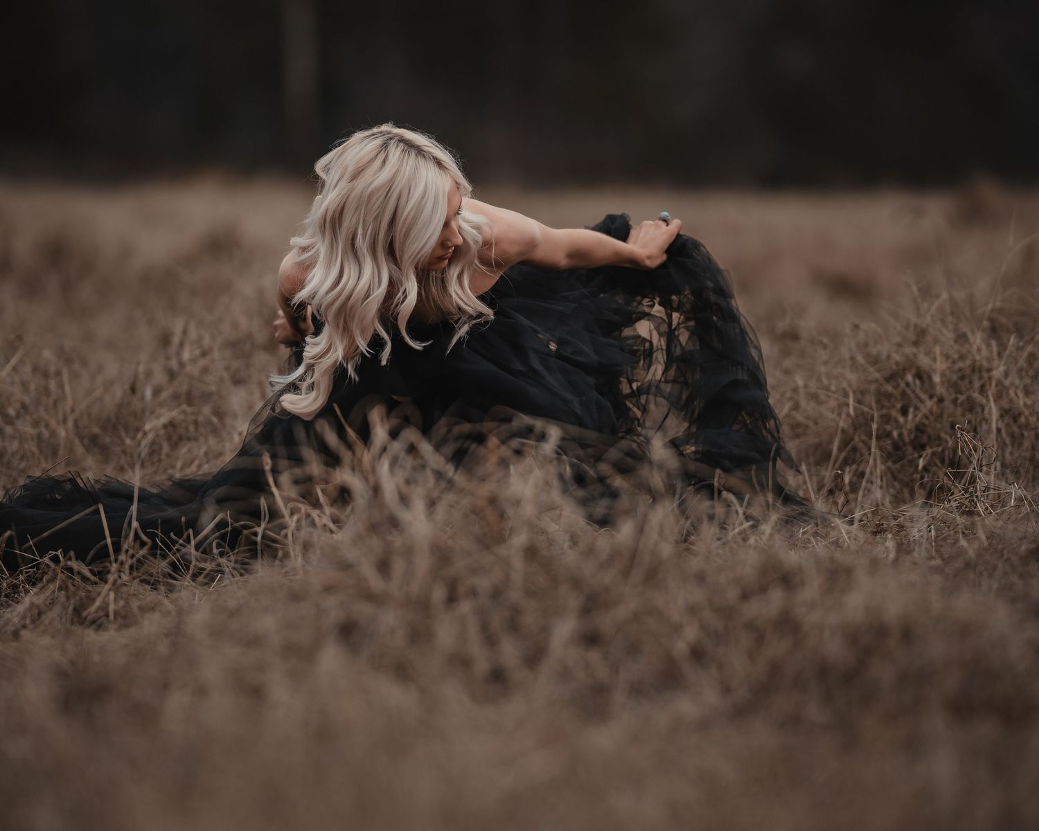 Woman in black gown sitting gracefully in a dried grass field.