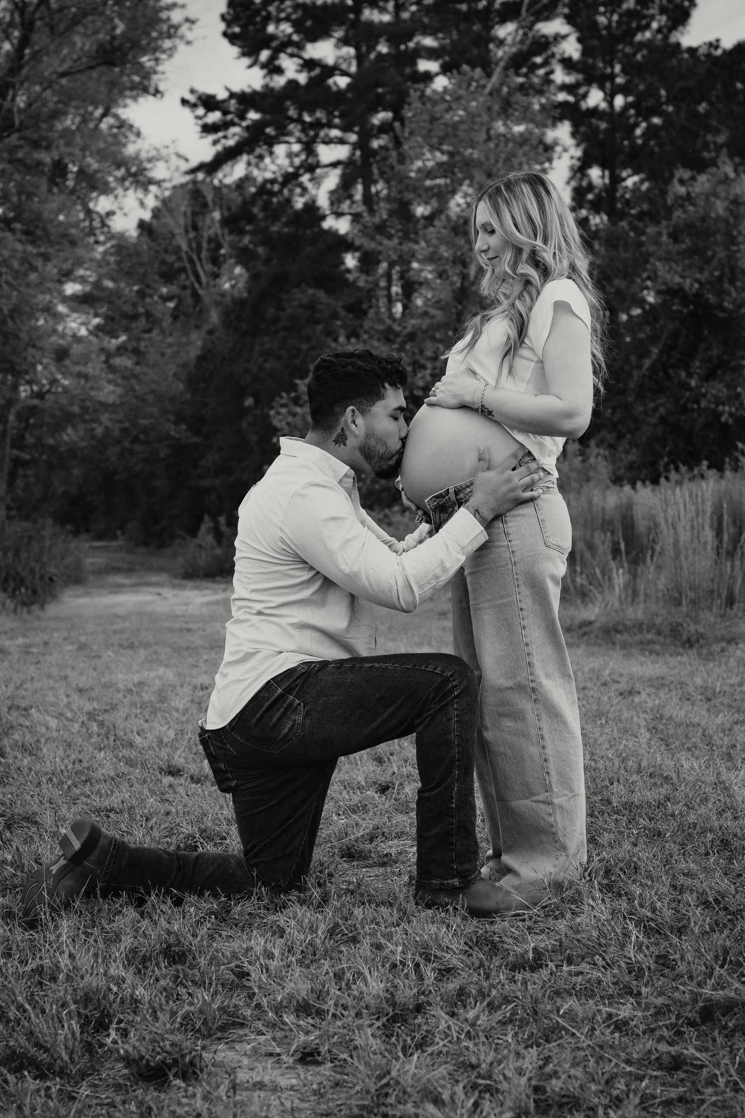 A tender black and white maternity photo captured outdoors with a partner kneeling in a grassy field.