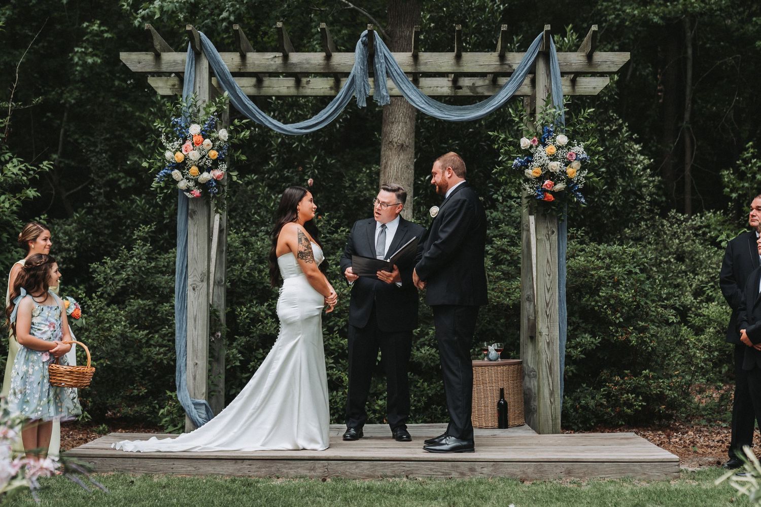 Couple exchanging vows at outdoor wedding ceremony under rustic wooden arbor with blue fabric and floral arrangements.