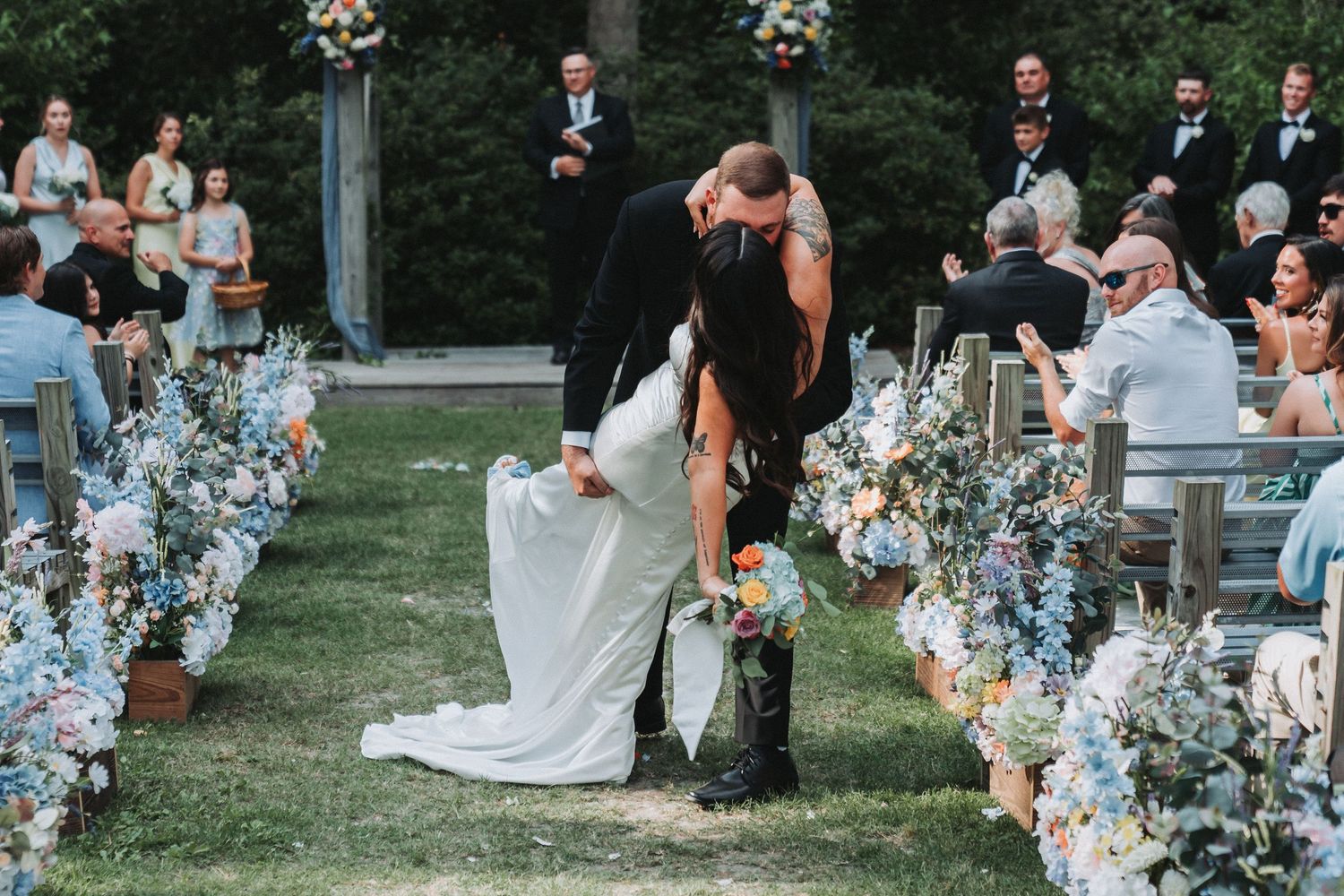 Bride and groom share a romantic dip kiss at outdoor wedding ceremony lined with blue and white floral arrangements.