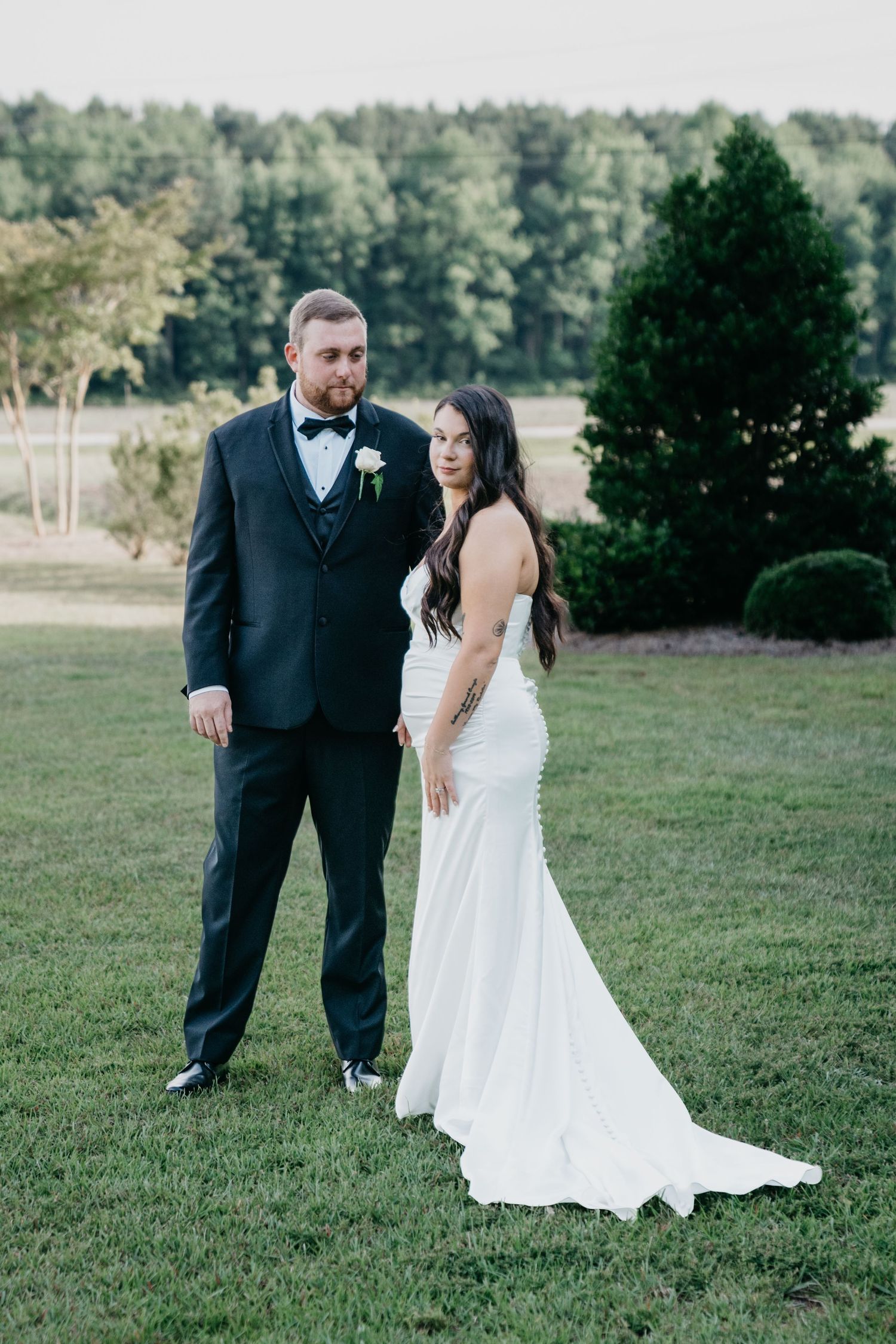 Bride in white gown and groom in black tuxedo pose together on green lawn with trees in background.