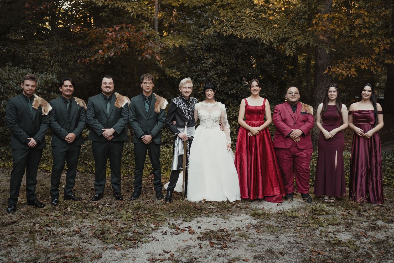 Wedding party in formal attire stands together outdoors with groomsmen in dark suits and bridesmaids in burgundy dresses.