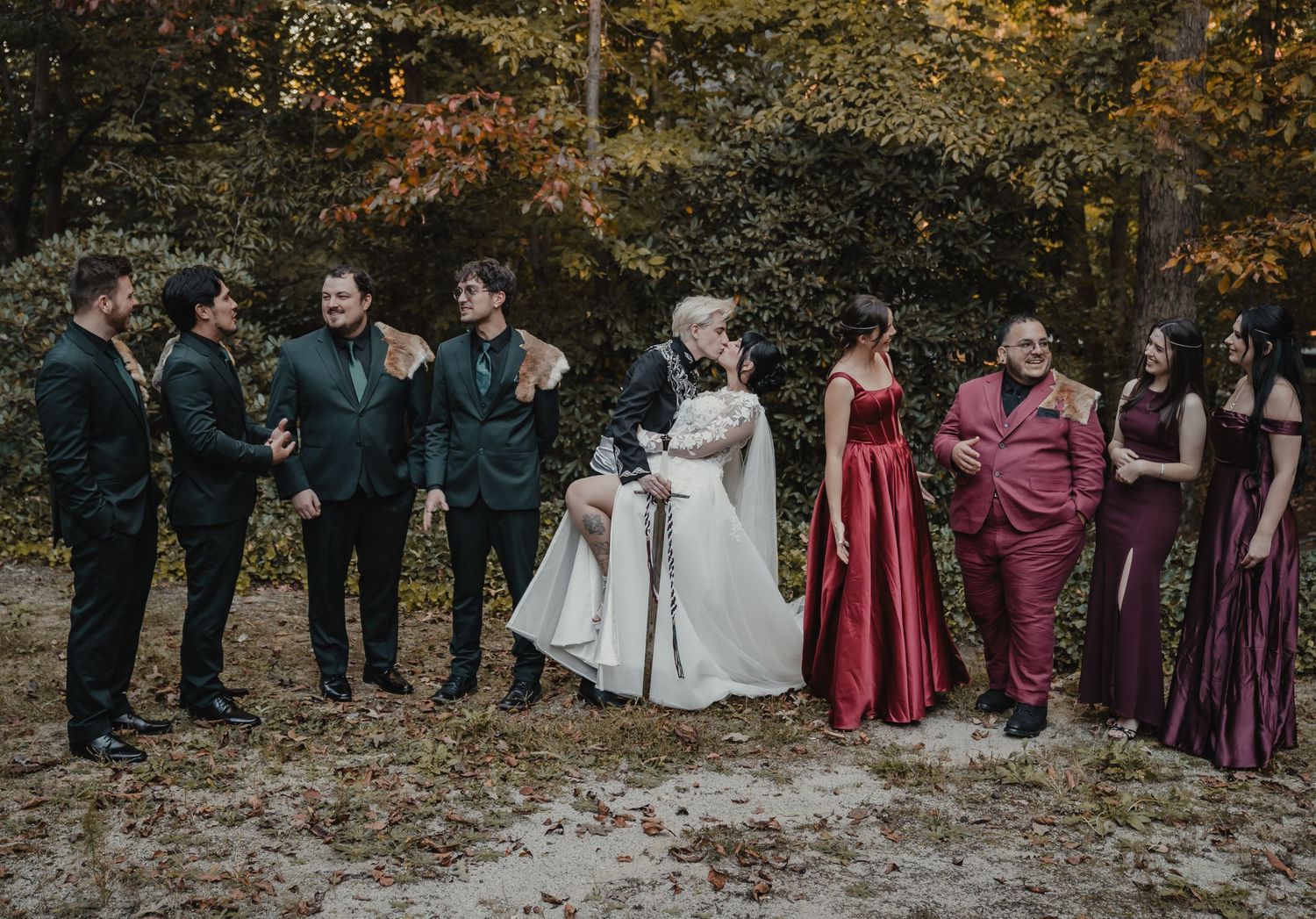 Wedding party in burgundy dresses and dark suits poses together in autumn forest setting with fallen leaves.