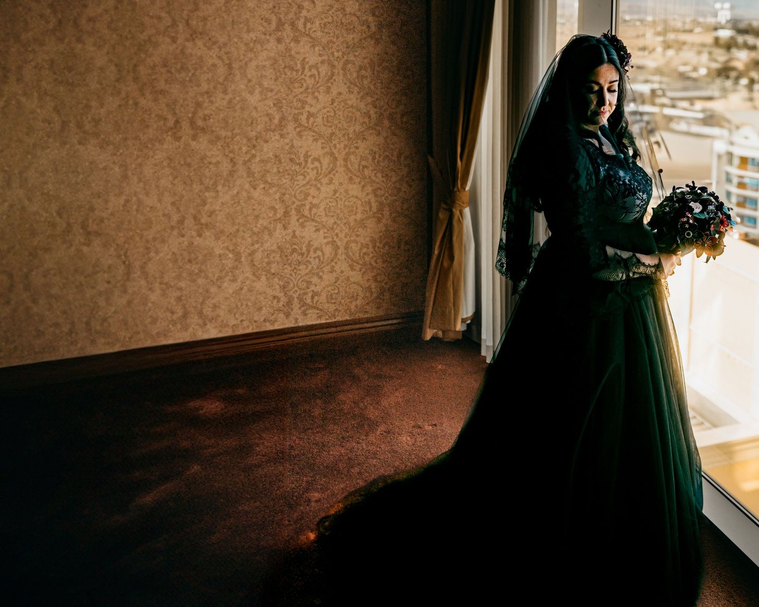 A person in an elegant dark green dress stands in a dimly lit room by a window.