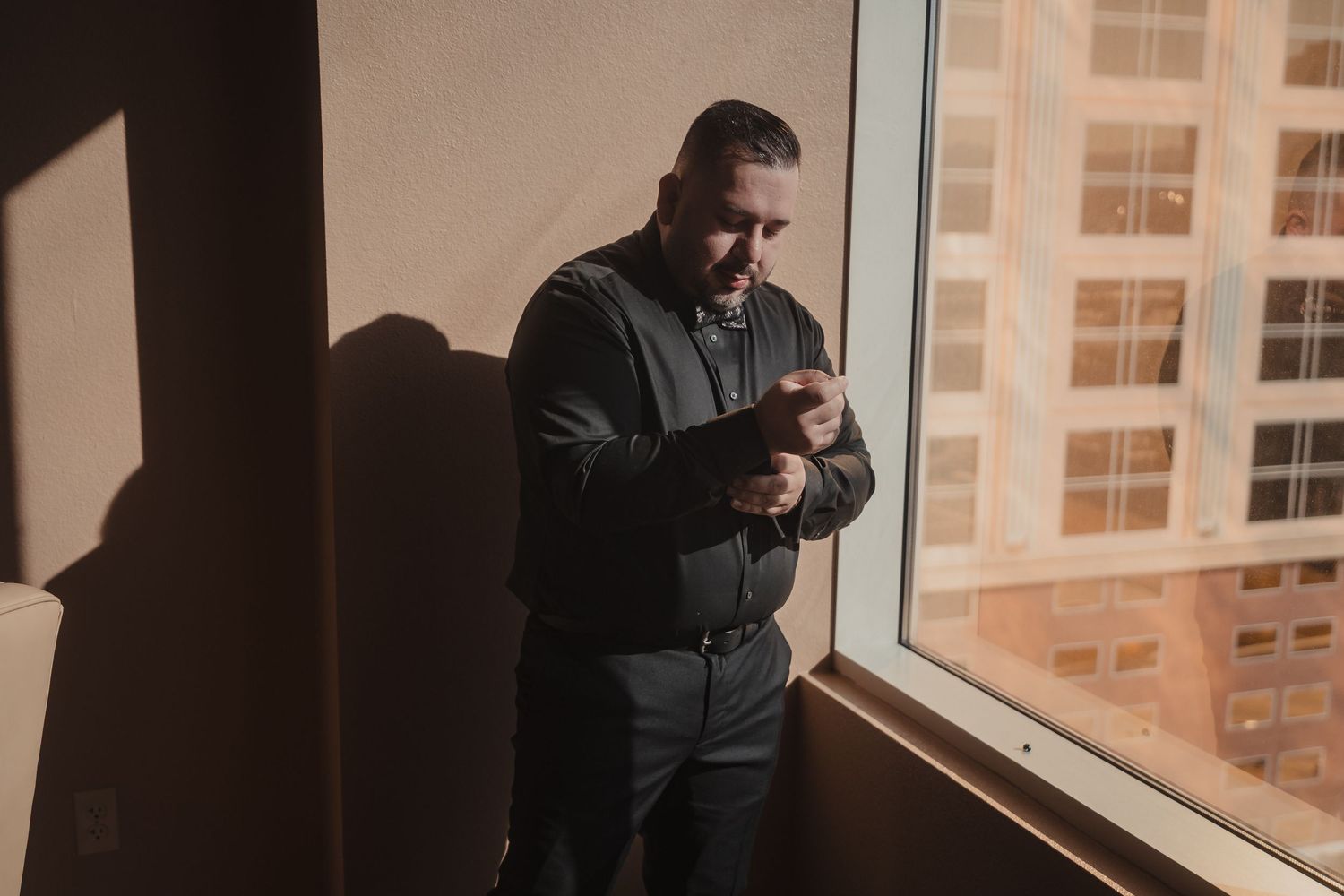 Person in dark clothing stands by window using mobile phone in dimly lit room.