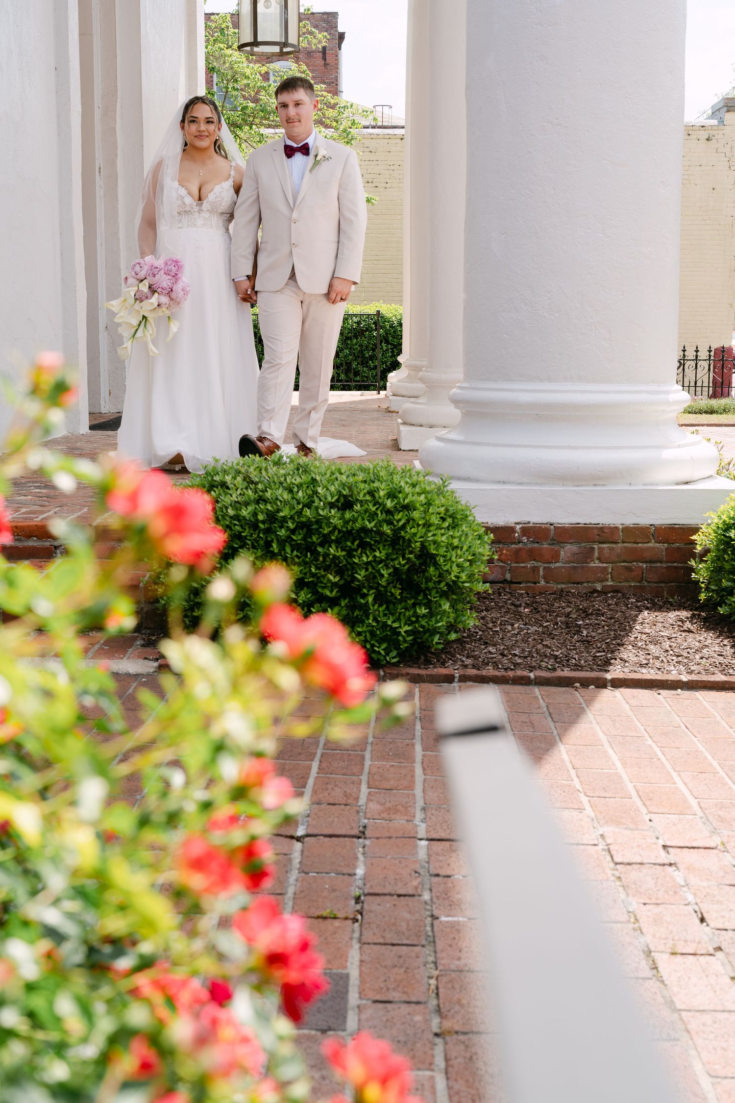 Bride and groom walking among white columns and blooming flowers, bride holding a bouquet of purple blooms.