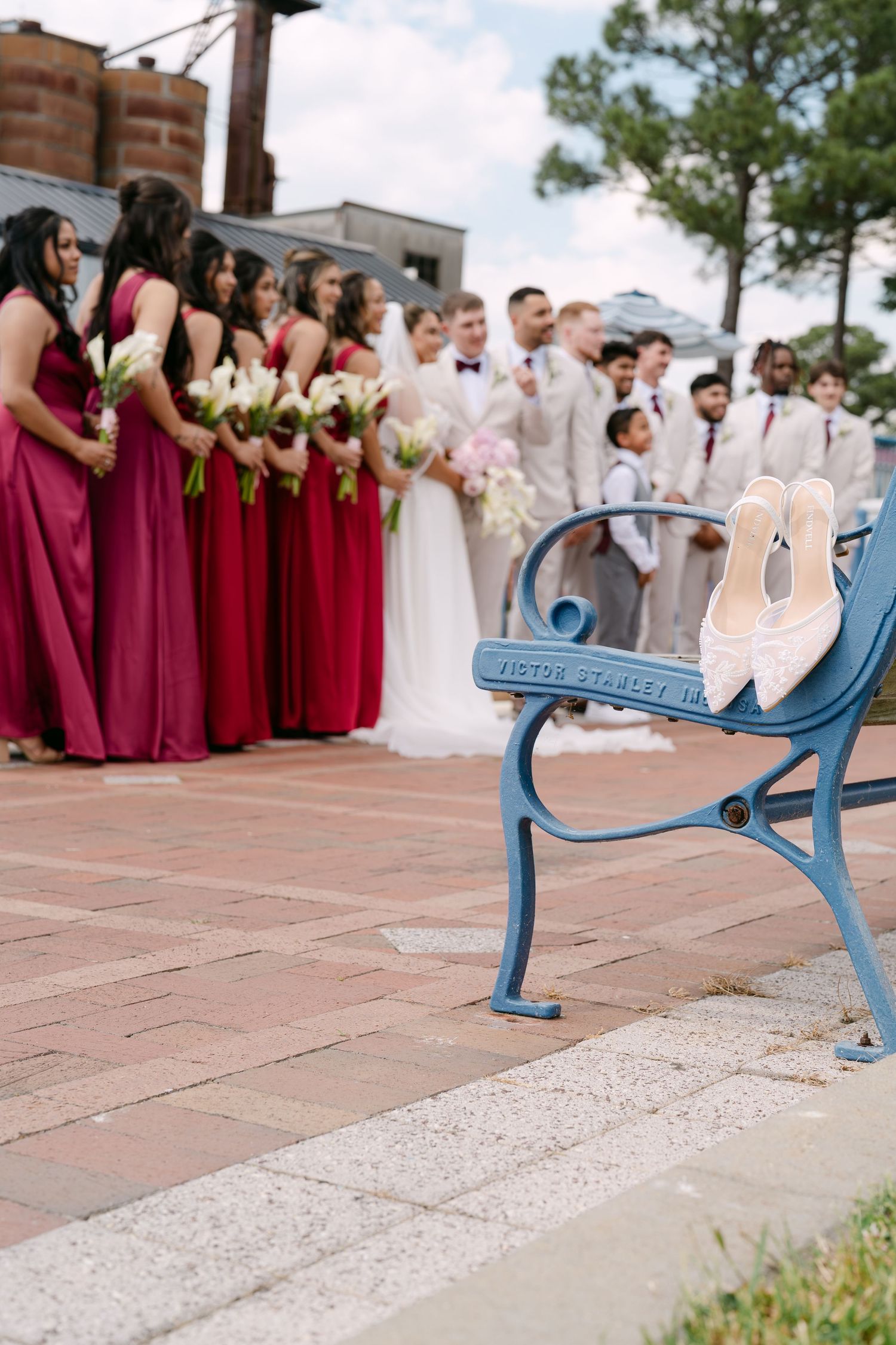 Wedding party in red and white attire poses outdoors while bridal heels rest on a blue bench in the foreground.