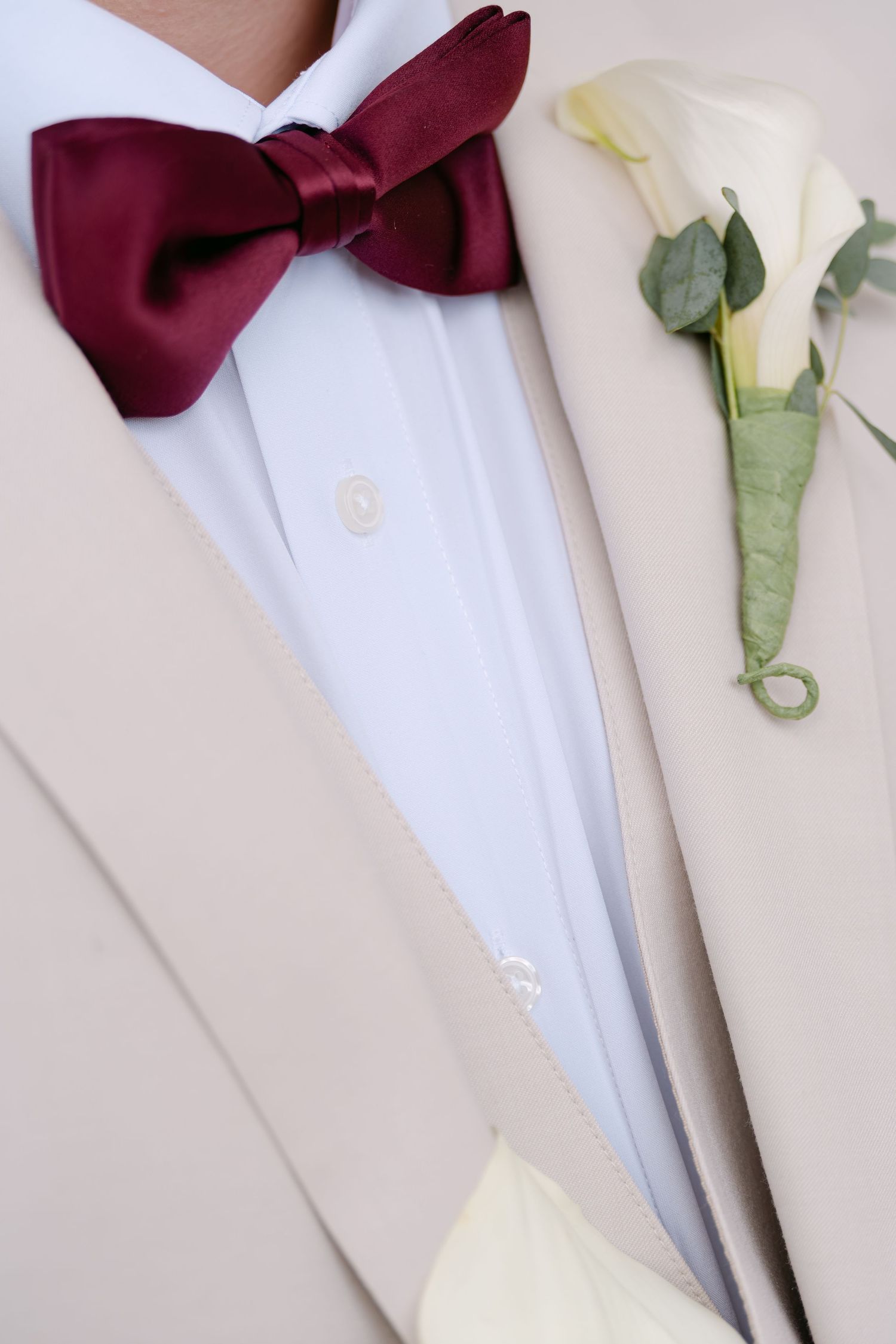Close-up of groom wearing cream tuxedo, burgundy bow tie, and white calla lily boutonniere on lapel.
