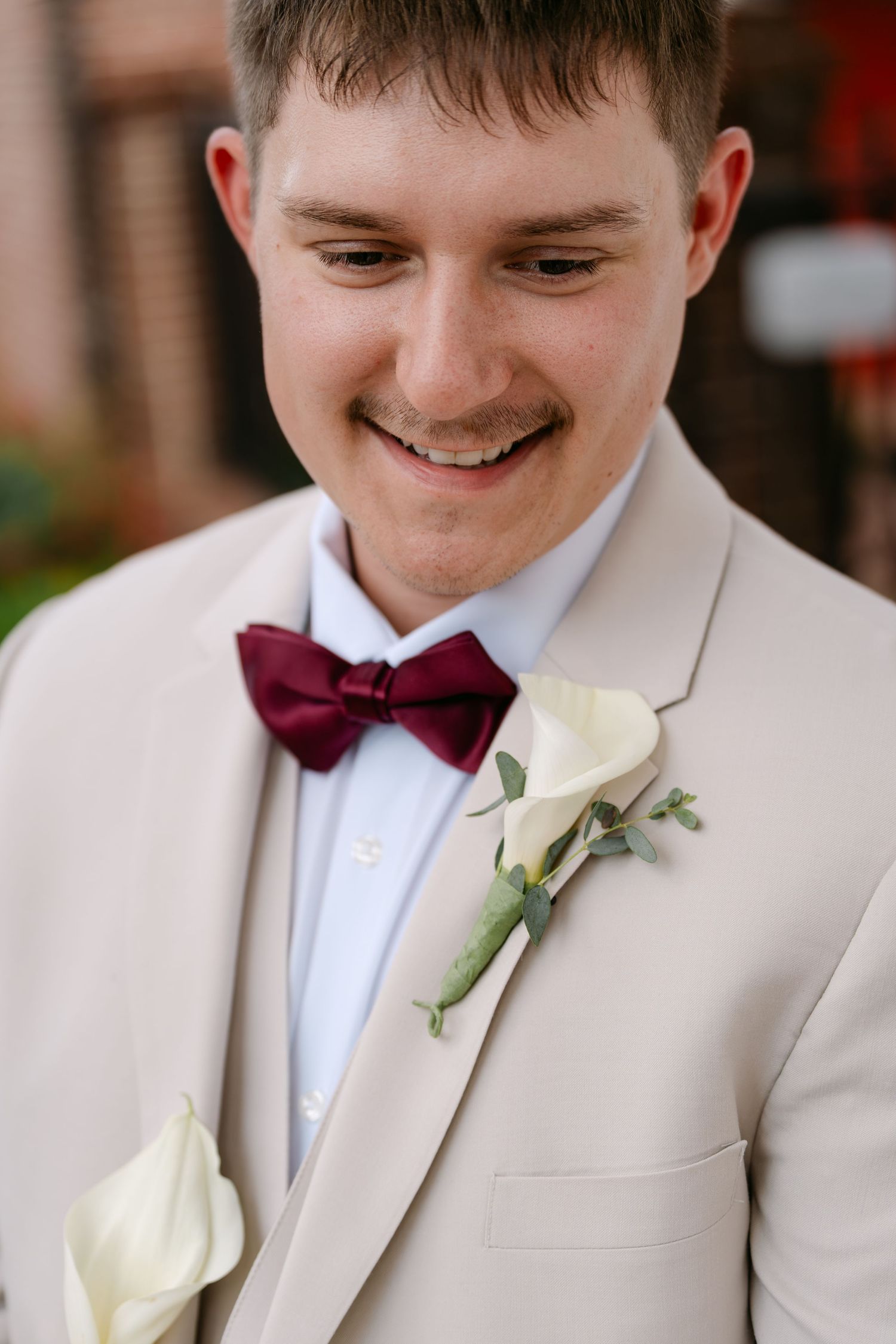 Smiling groom in beige suit with burgundy bow tie and white calla lily boutonniere.
