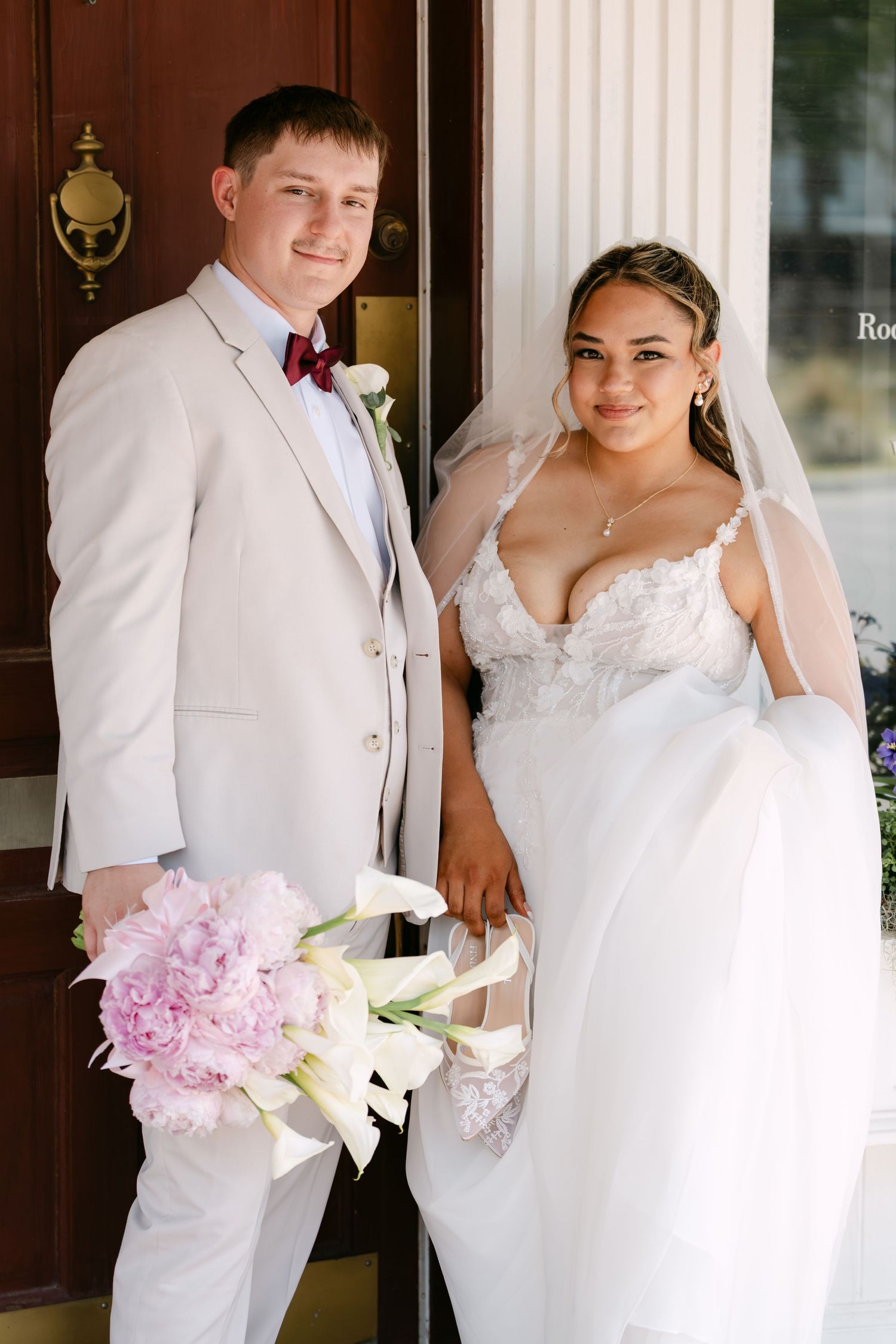 Bride and groom posing together, she in white lace gown with veil, he in beige suit with burgundy bow tie.