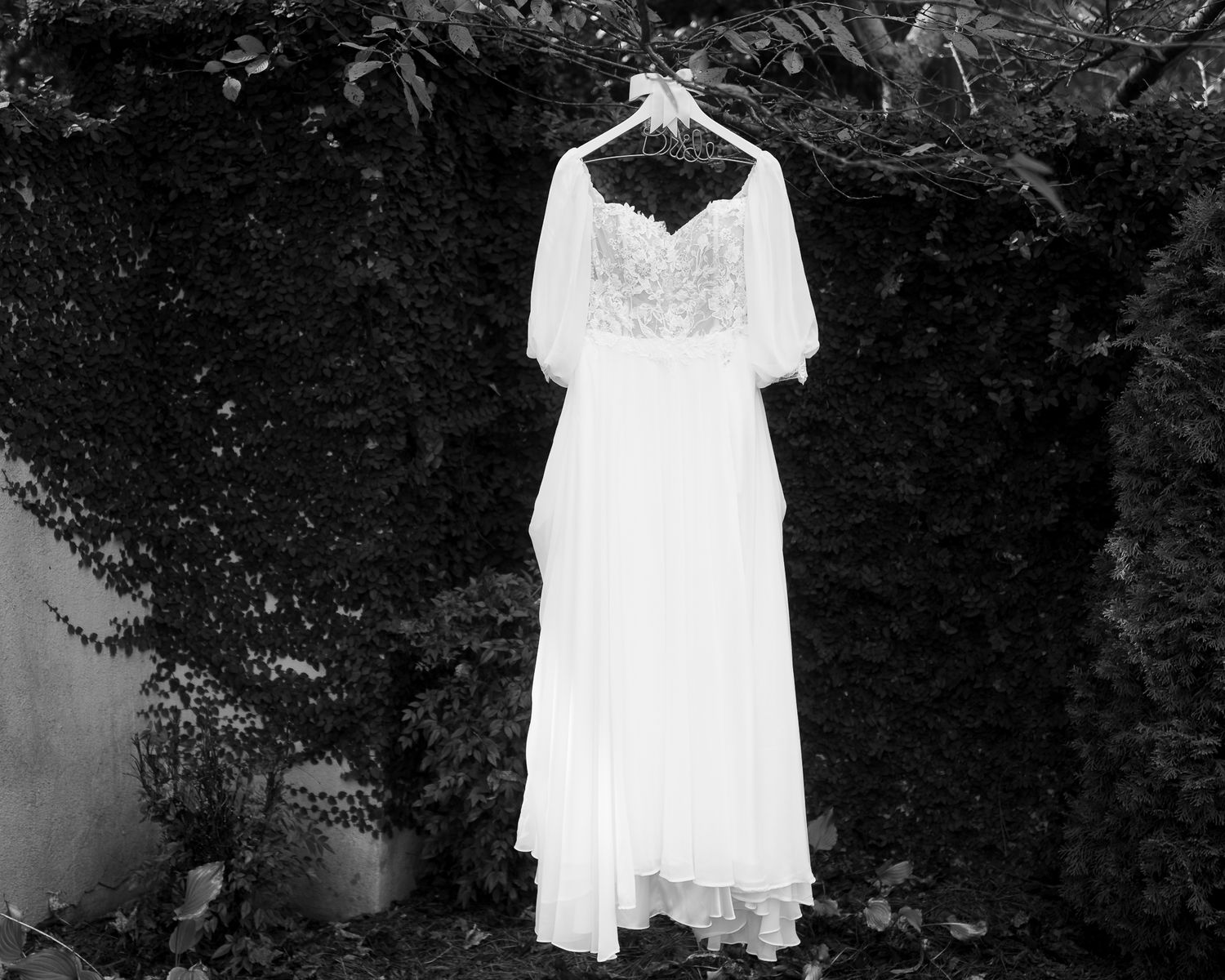 White lace wedding dress hanging outdoors against dark foliage creating ethereal contrast in black and white photo.