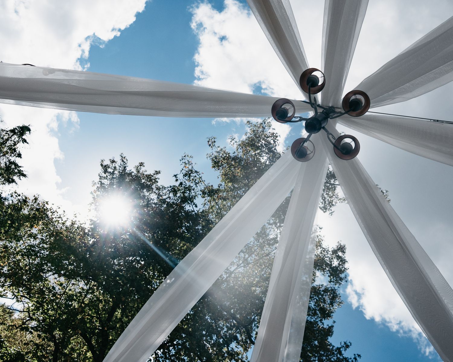 White fabric canopy panels create a geometric pattern against a blue sky with puffy clouds and green trees.