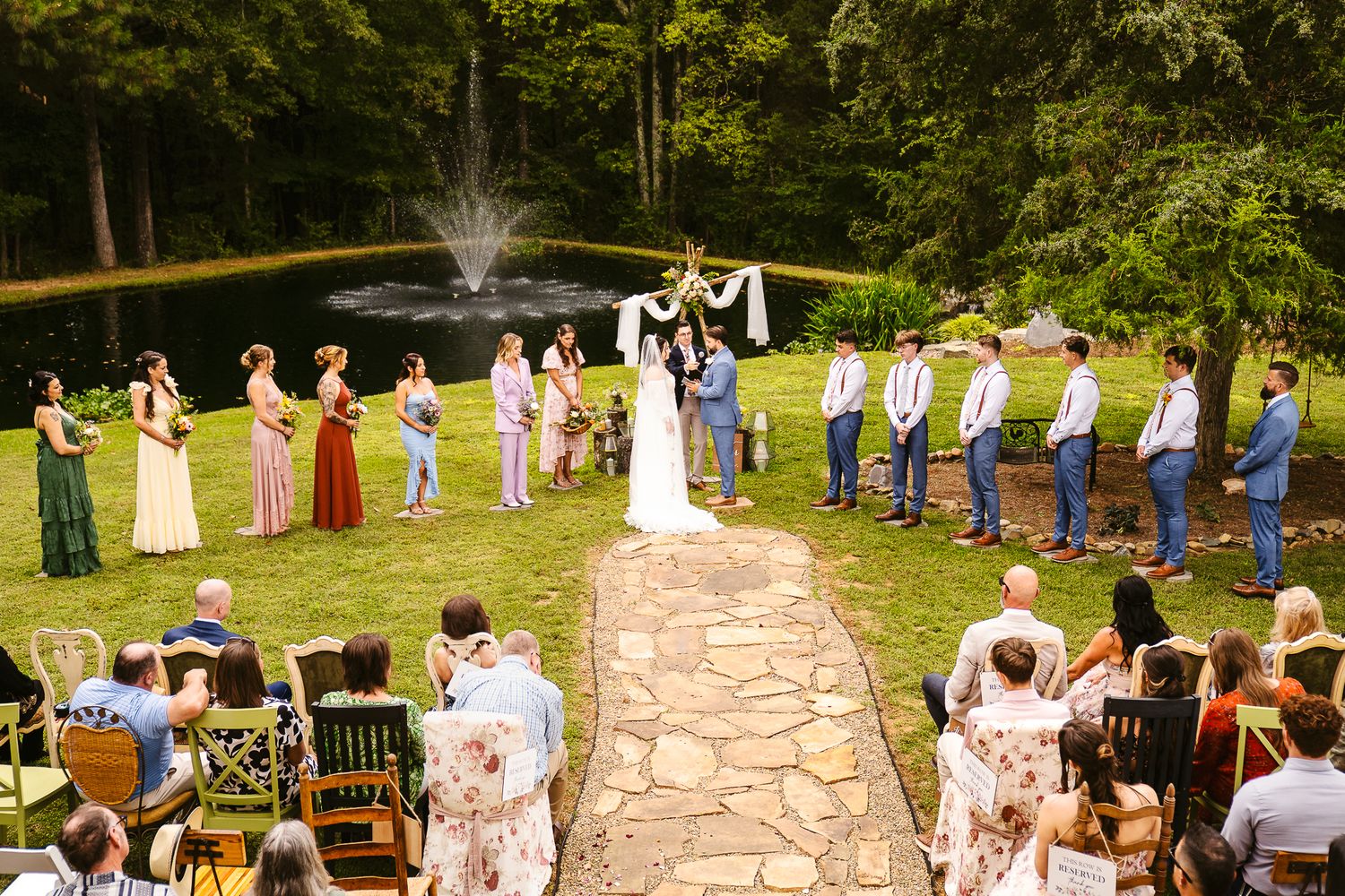 Wedding guests gather outdoors for a ceremony in a scenic garden setting with stone pathways and natural landscaping.